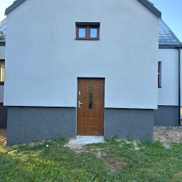 Nidecki Elewacje - Nowoczesny dom z jasną elewacją i ciemnoszarą podmurówką, brązowe drzwi wejściowe z przeszkleniami, małe okno nad drzwiami, trawnik przed budynkiem.