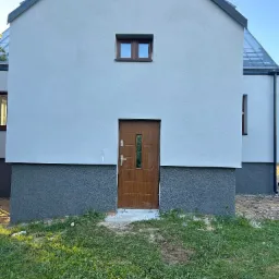 Nowoczesny dom z jasną elewacją i ciemnoszarą podmurówką, brązowe drzwi wejściowe z przeszkleniami, małe okno nad drzwiami, trawnik przed budynkiem.