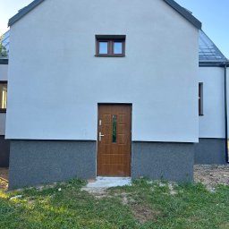 Nidecki Elewacje - Nowo wykończona elewacja domu jednorodzinnego z brązowymi drzwiami wejściowymi i małym oknem na górze, dolna część ściany wykończona ciemniejszym tynkiem strukturalnym.