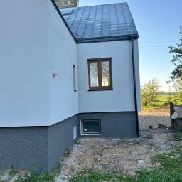 Nidecki Elewacje - Fragment elewacji domu po tynkowaniu, z widocznym podziałem na część dolną w ciemniejszym, ziarnistym tynku i górną w jasnym, gładkim tynku, okna z brązowymi ramami, metalowy dach i antena...