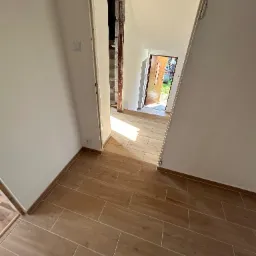Wnętrze domu w trakcie remontu: widok na korytarz z nową podłogą z płytek imitujących drewno, ściany przygotowane do malowania, widoczne gniazdko elektryczne i fragment nieotynkowanego przejścia...