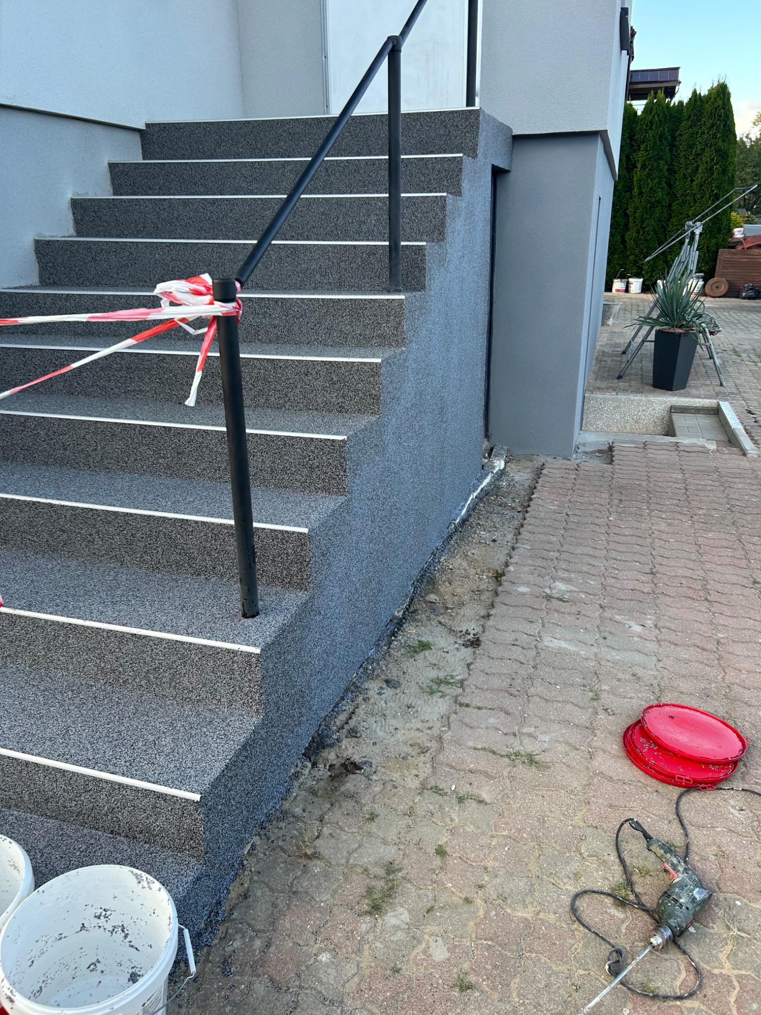 Schody wejściowe do domu pokryte szarą żywicą epoksydową z białymi listwami, zabezpieczone taśmą ostrzegawczą, widoczne wiaderka i mieszadło elektryczne na kostce brukowej.