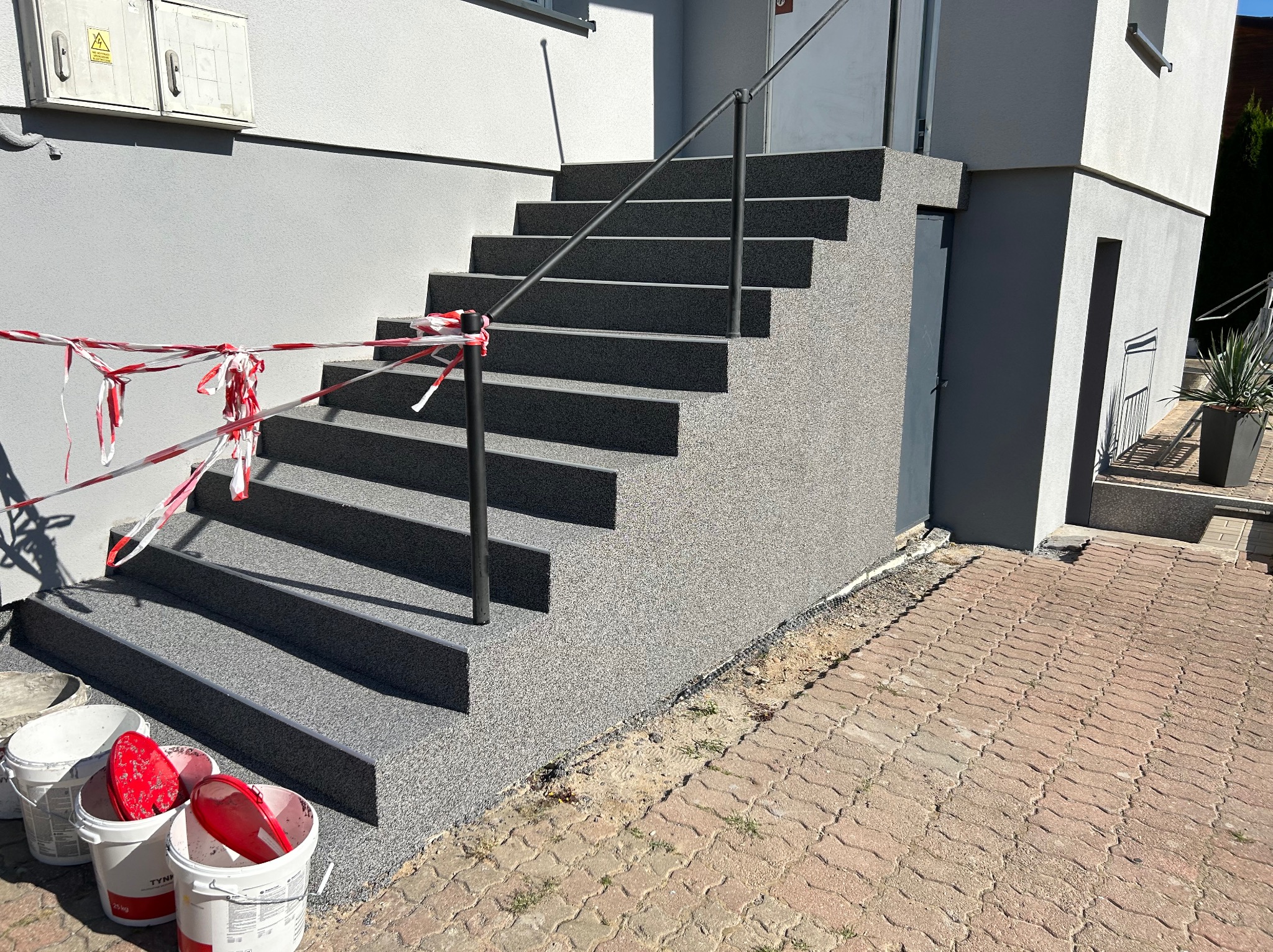 Schody wejściowe do budynku po nałożeniu szarego tynku mozaikowego, zabezpieczone taśmą ostrzegawczą, widoczne wiadra z resztkami tynku.