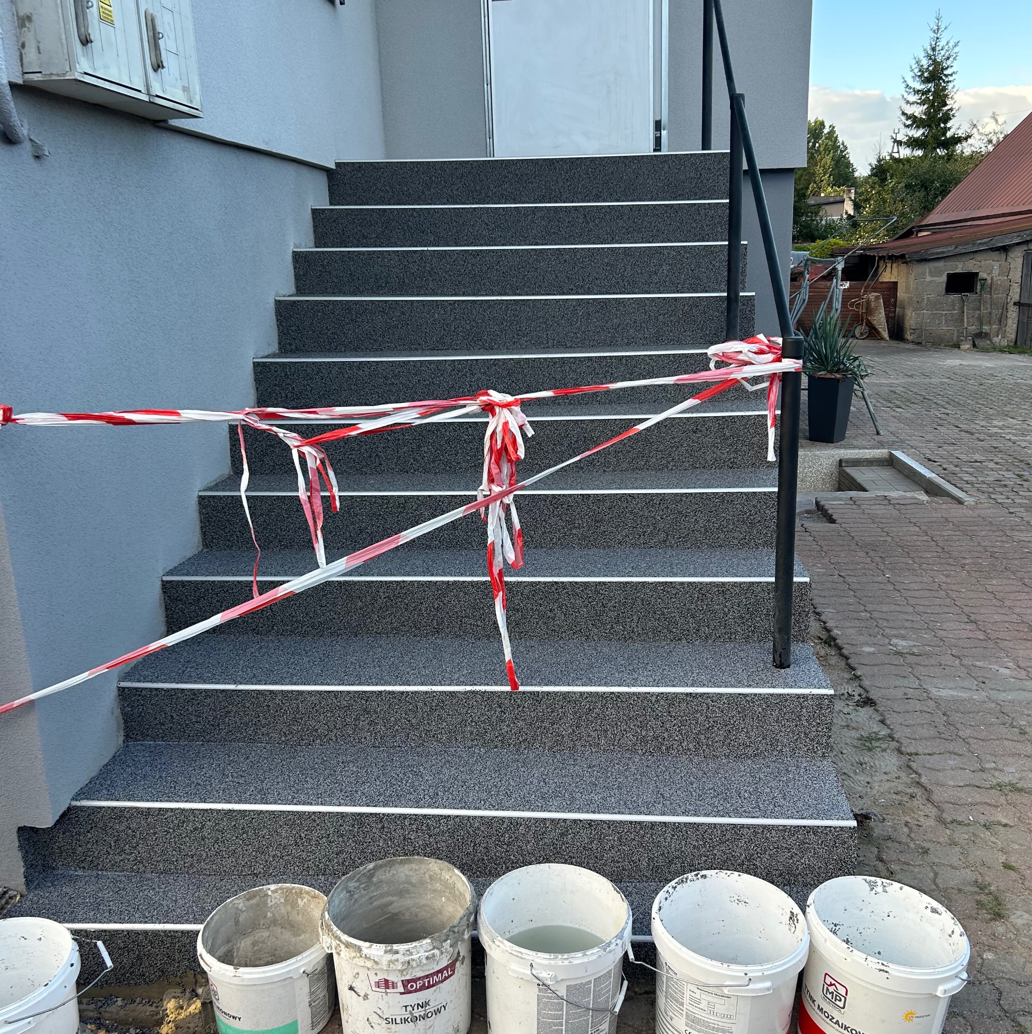 Schody wejściowe do domu po renowacji, zabezpieczone taśmą ostrzegawczą, widoczne wiadra z resztkami materiałów budowlanych, elewacja budynku w odcieniach szarości.
