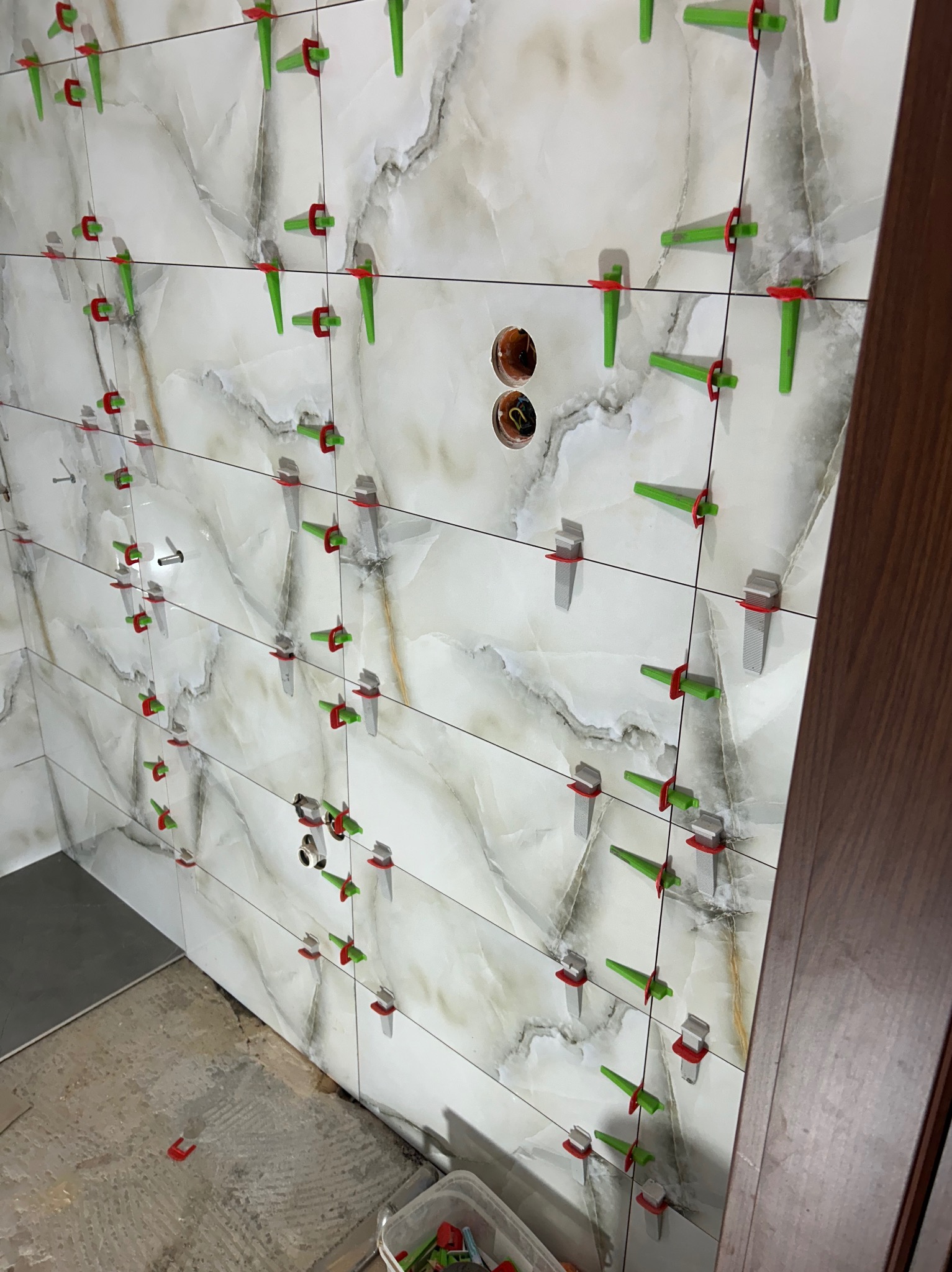 Układanie marmurkowych płytek ceramicznych na ścianie z użyciem systemu poziomowania płytek (klipsy i kliny), widoczne otwory na gniazdka elektryczne i fragment podłogi przed fugowaniem.