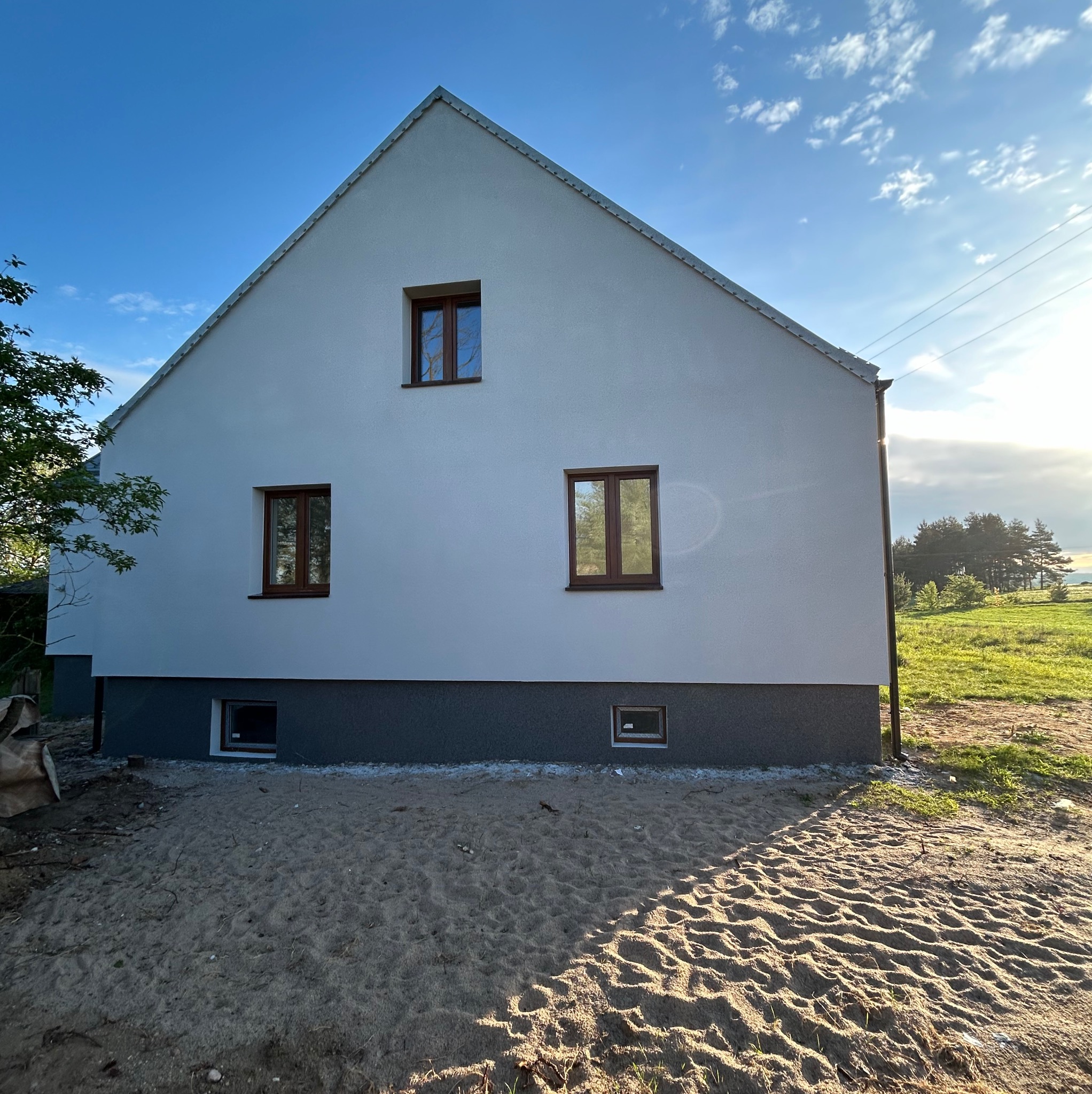 Boczny widok domu z nową, jasną elewacją i ciemną podmurówką, drewniane okna, piasek wokół fundamentów w słoneczny dzień.