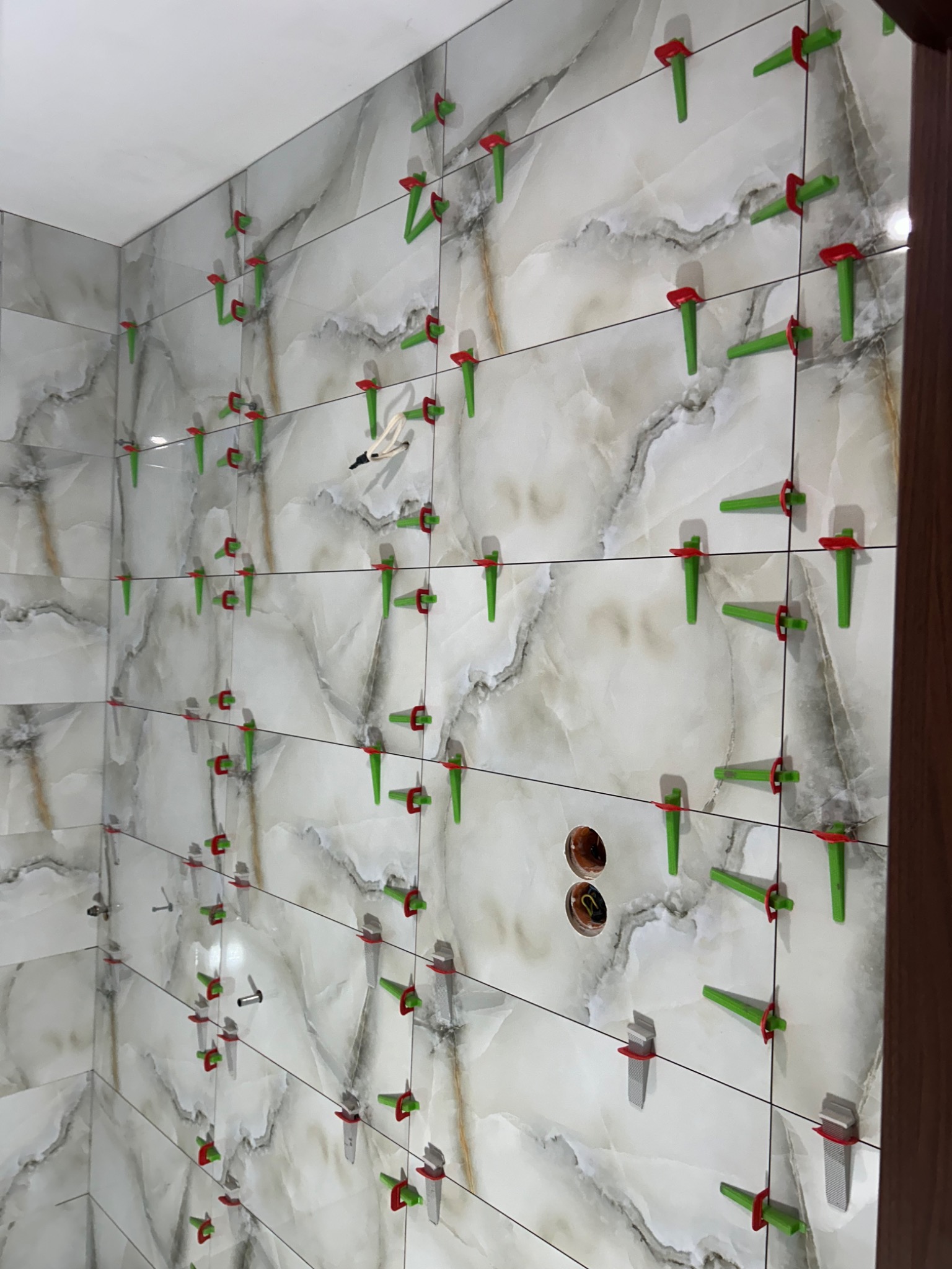 Ściana w trakcie układania płytek imitujących marmur, z użyciem poziomujących klipsów z czerwonymi klinami, widoczne otwory pod gniazdka elektryczne.