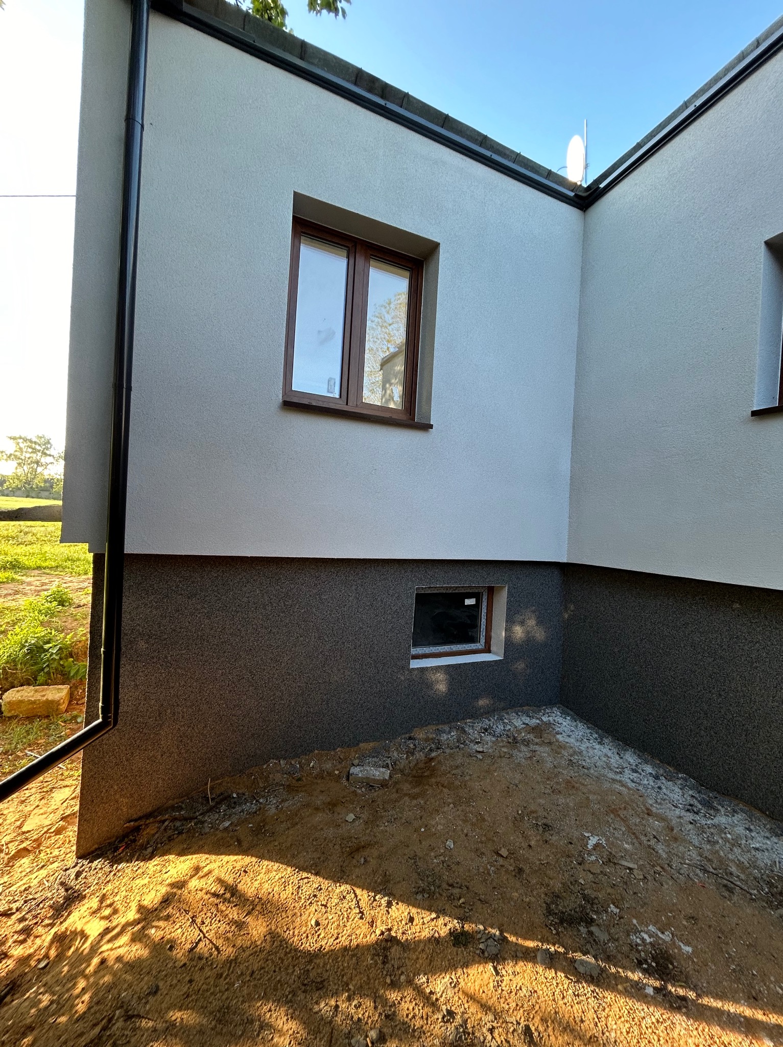 Fragment domu z ocieploną elewacją, widoczna dwukolorowa struktura tynku, okna z brązowymi ramami i rynna spustowa przy narożniku budynku, teren wokół budynku częściowo przygotowany...