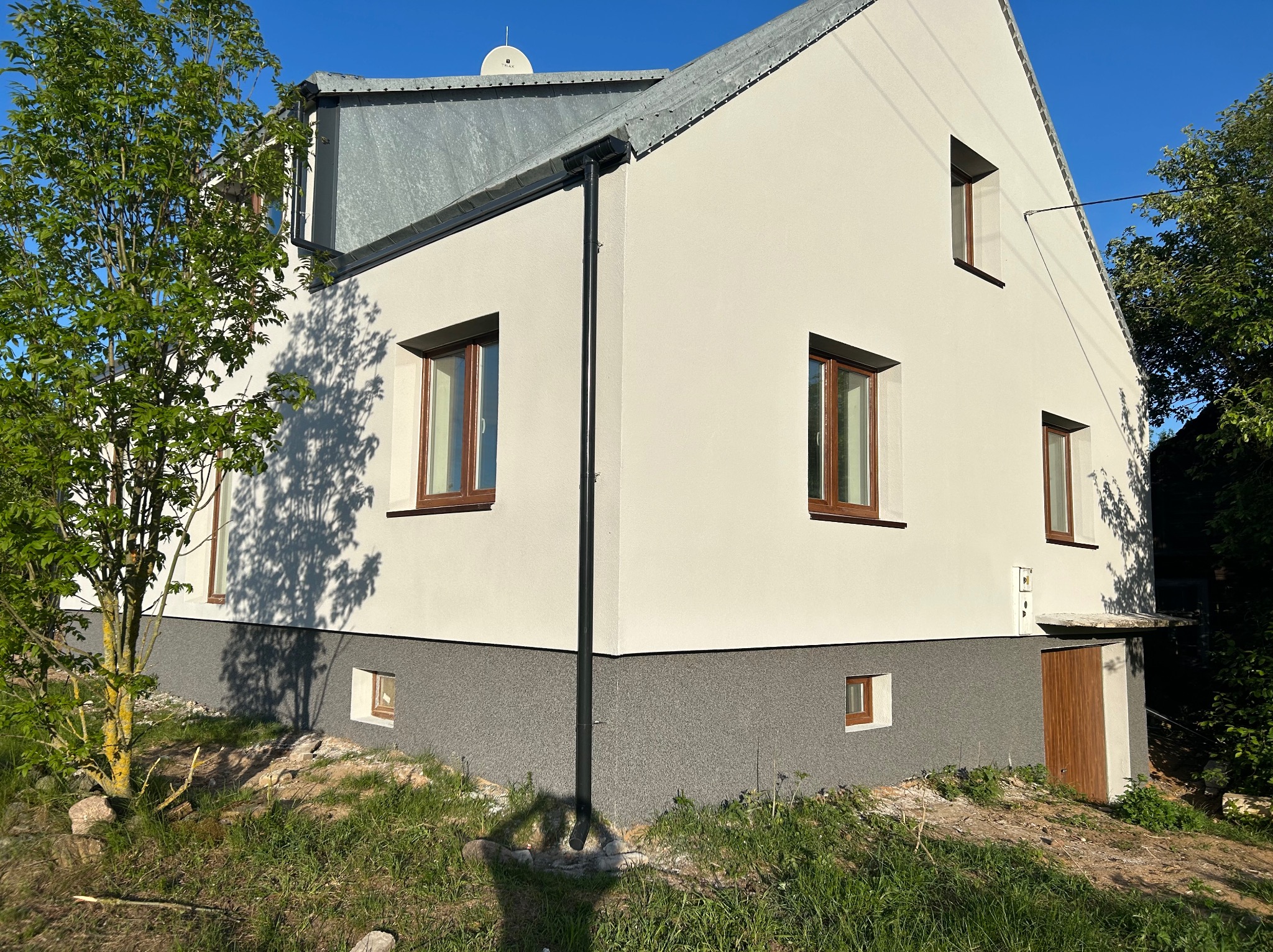 Dwukondygnacyjny dom jednorodzinny z jasną elewacją i szarą podmurówką, brązowe ramy okienne, metalowy dach, widoczny fragment ogrodu i cień drzewa.