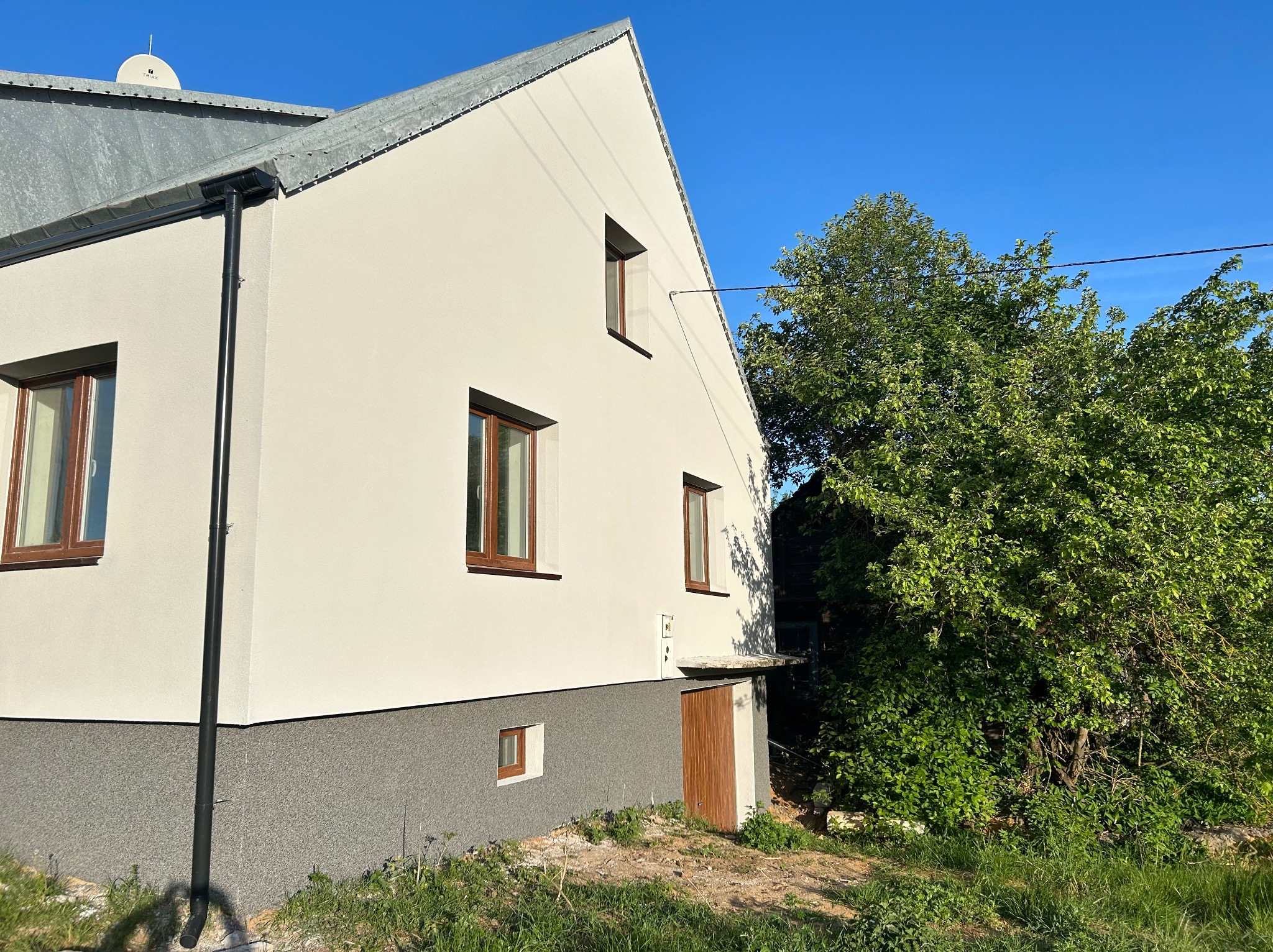 Świeżo otynkowana ściana domu z brązowymi oknami, dolna część ściany w szarym kolorze, widok z boku na tle błękitnego nieba i zieleni drzew.