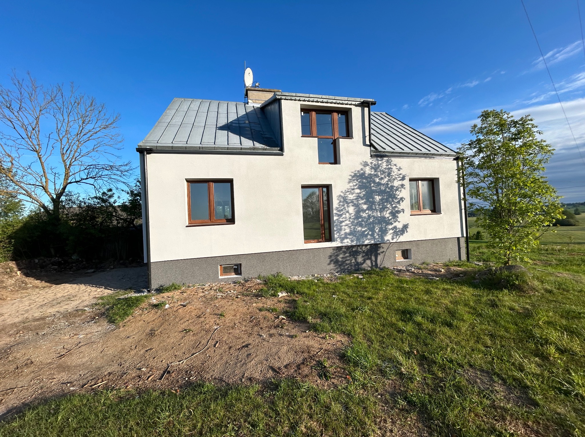 Nowoczesny dom z białą elewacją i szarym dachem blachodachówką, widoczny z poziomu gruntu, z fragmentem niezagospodarowanego terenu wokół, w słoneczny dzień.