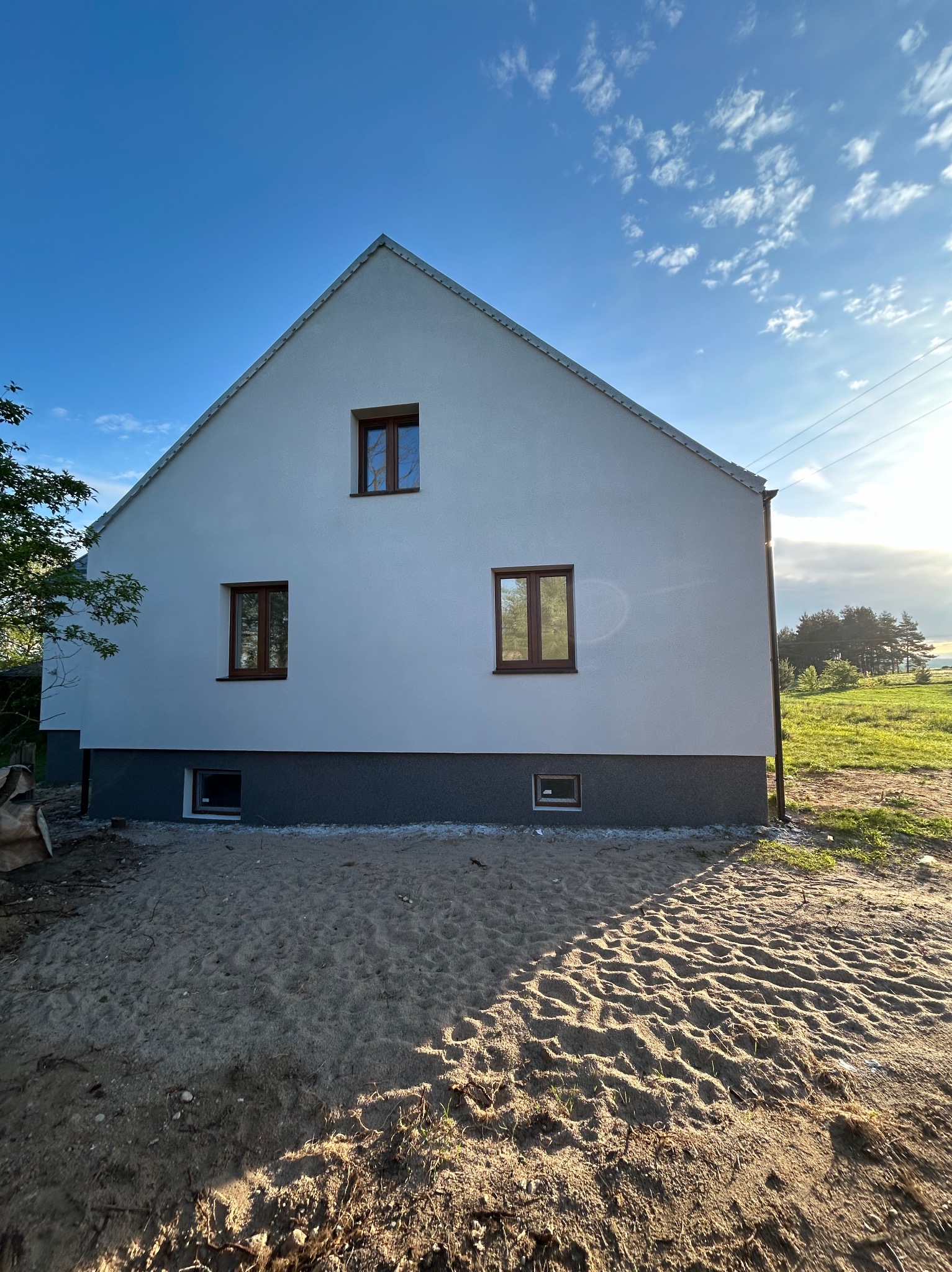 Świeżo otynkowana, biała elewacja domu jednorodzinnego z brązowymi oknami, widok z poziomu gruntu na tle błękitnego nieba z chmurami.