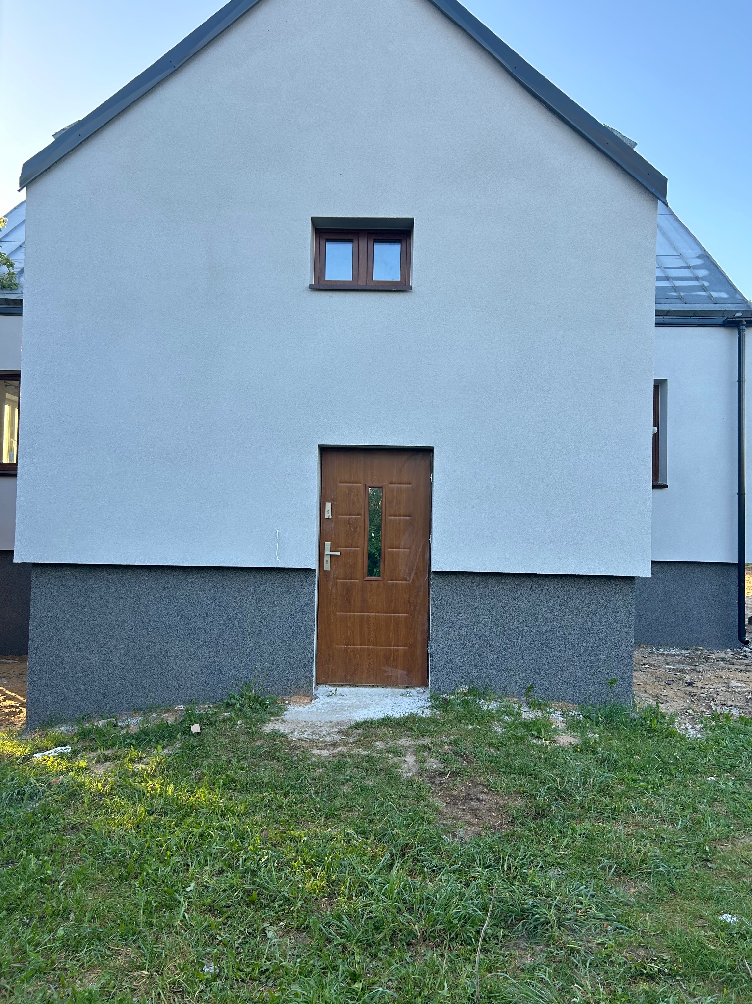 Nowoczesny dom z jasną elewacją i ciemnoszarą podmurówką, brązowe drzwi wejściowe z przeszkleniami, małe okno nad drzwiami, trawnik przed budynkiem.