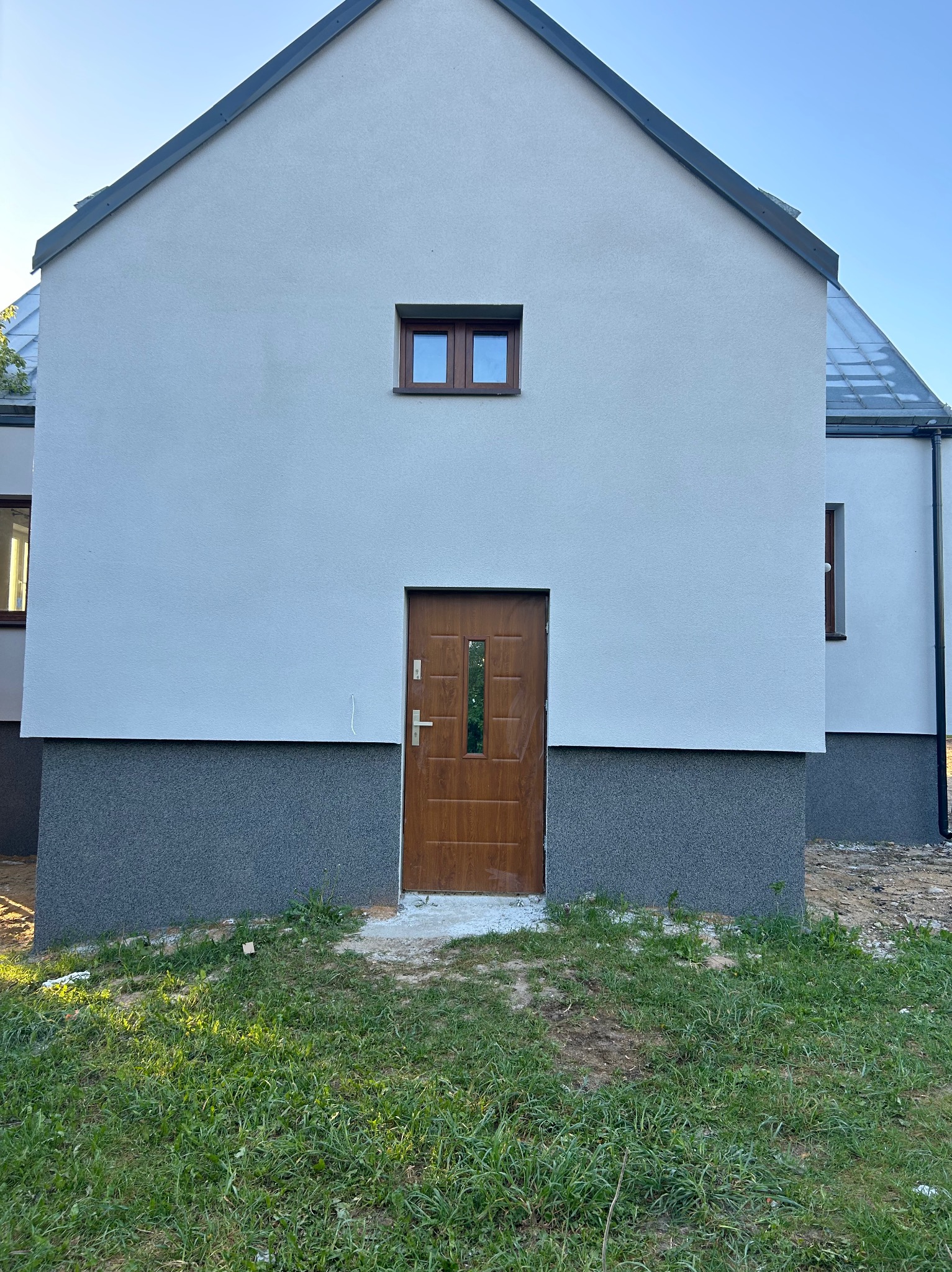 Nowo wykończona elewacja domu jednorodzinnego z brązowymi drzwiami wejściowymi i małym oknem na górze, dolna część ściany wykończona ciemniejszym tynkiem strukturalnym.