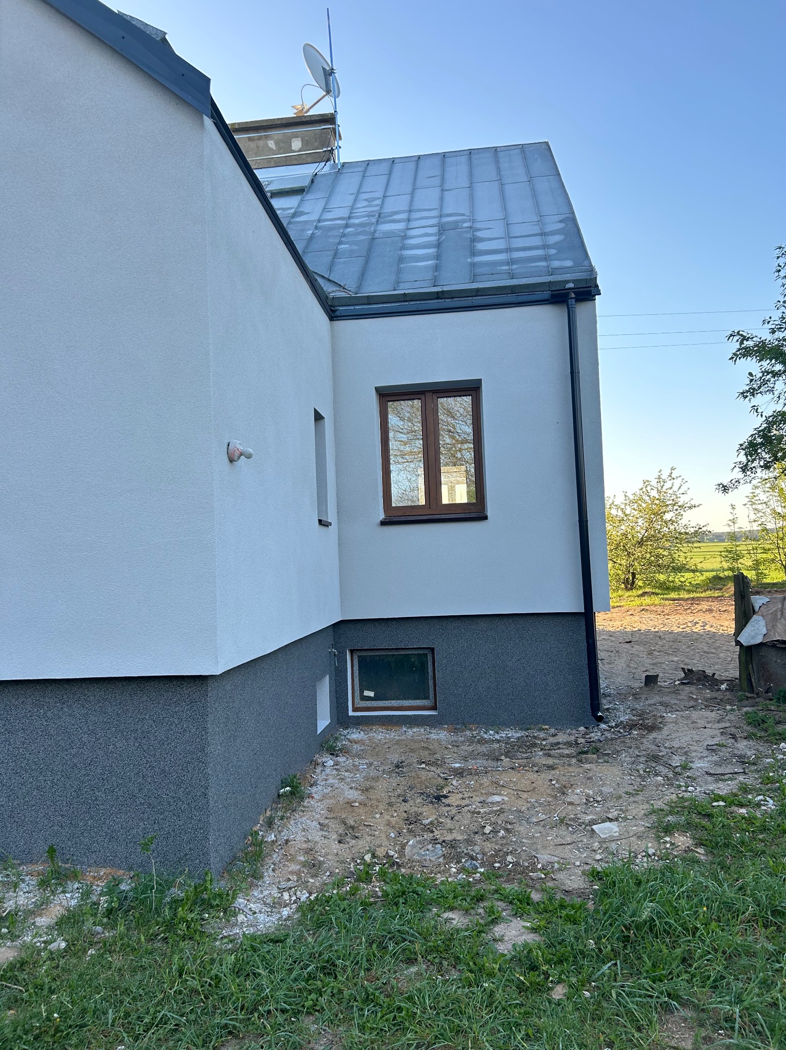 Fragment elewacji domu po tynkowaniu, z widocznym podziałem na część dolną w ciemniejszym, ziarnistym tynku i górną w jasnym, gładkim tynku, okna z brązowymi ramami, metalowy dach i antena...