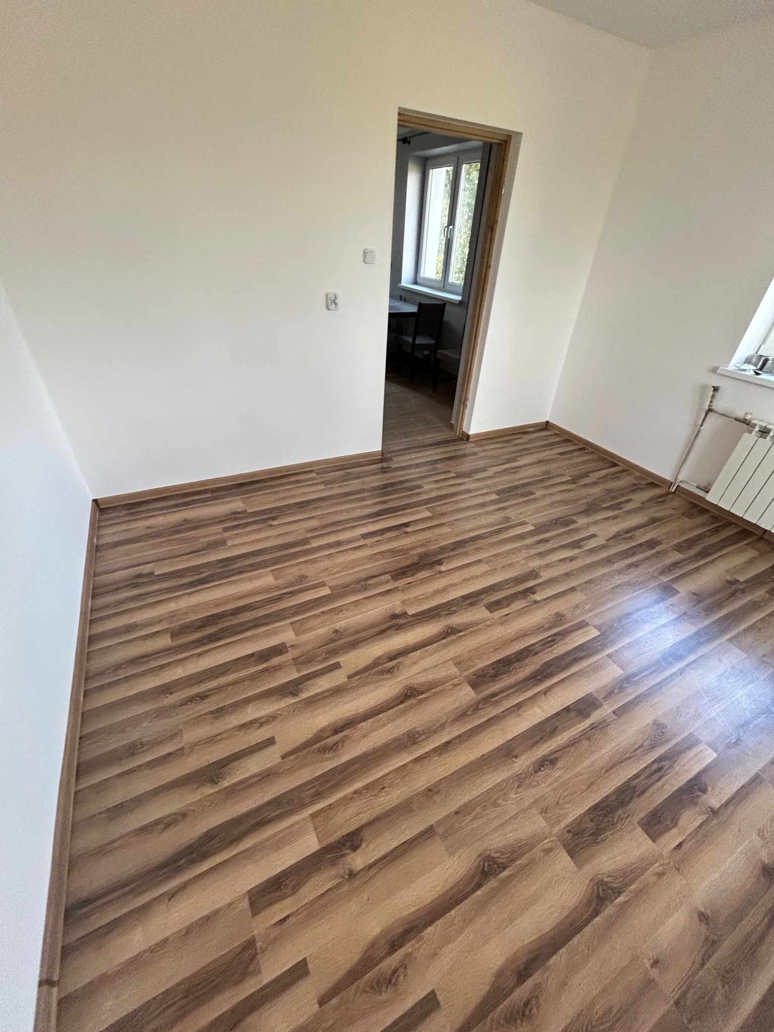 Pusty pokój z nową podłogą z paneli imitujących drewno, białe ściany, grzejnik pod oknem i widok na sąsiedni pokój przez otwarte drzwi.