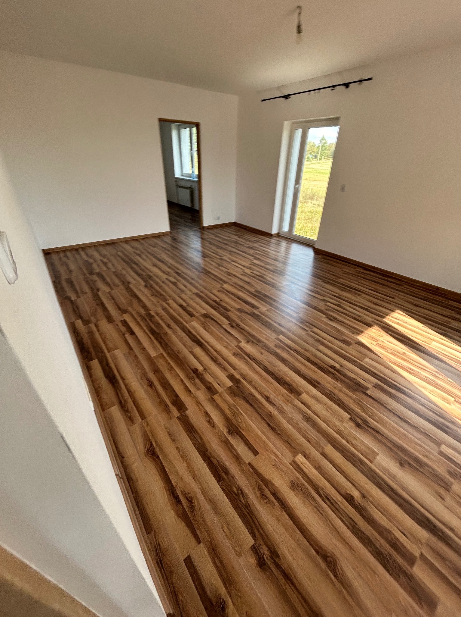 Puste, jasne pomieszczenie z panelami podłogowymi imitującymi drewno, widok na okno i drzwi balkonowe, ściany pomalowane na biało, listwy przypodłogowe w kolorze podłogi.