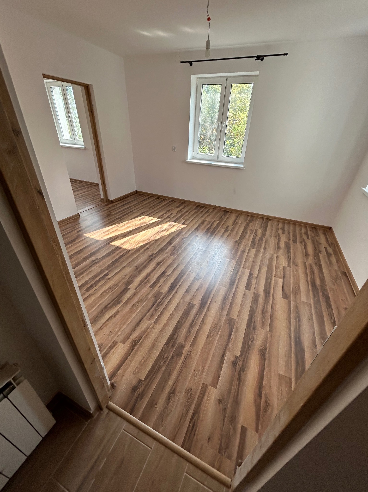 Pusty pokój z nową podłogą laminowaną w odcieniach brązu, wpadające promienie słoneczne przez okno, świeżo pomalowane białe ściany, widok z korytarza.