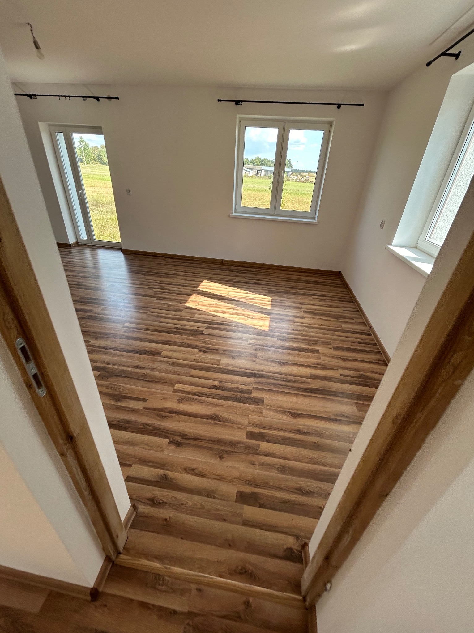 Pusty pokój z drewnianą podłogą, widok zza drewnianej framugi drzwi, okna wpuszczają naturalne światło, widok na zewnątrz na pole i zabudowania.