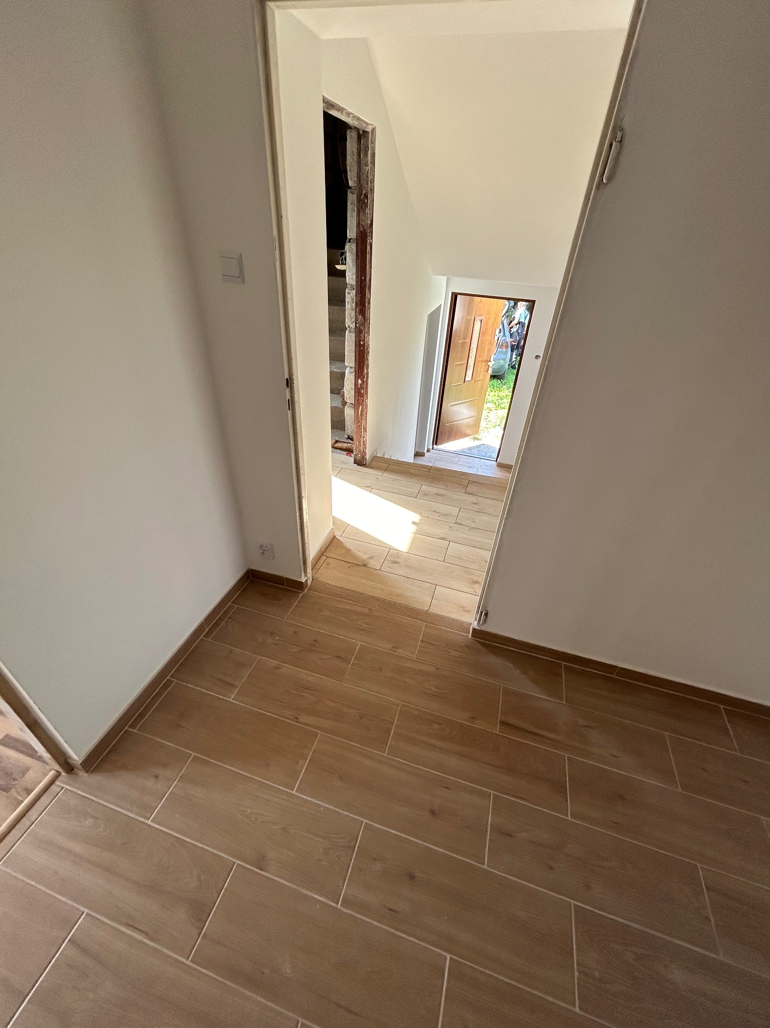 Wnętrze domu w trakcie remontu: widok na korytarz z nową podłogą z płytek imitujących drewno, ściany przygotowane do malowania, widoczne gniazdko elektryczne i fragment nieotynkowanego przejścia...