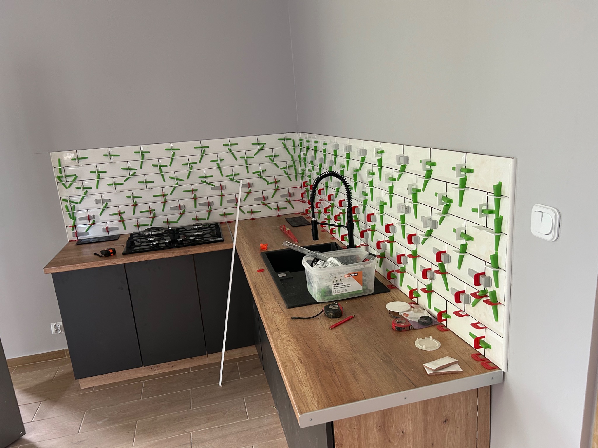 Montaż białych płytek w kuchni z użyciem klipsów poziomujących w kolorach zielonym, czerwonym i szarym. Widoczny fragment blatu z czarnym zlewem, bateria, płyta gazowa i narzędzia glazurnicze.