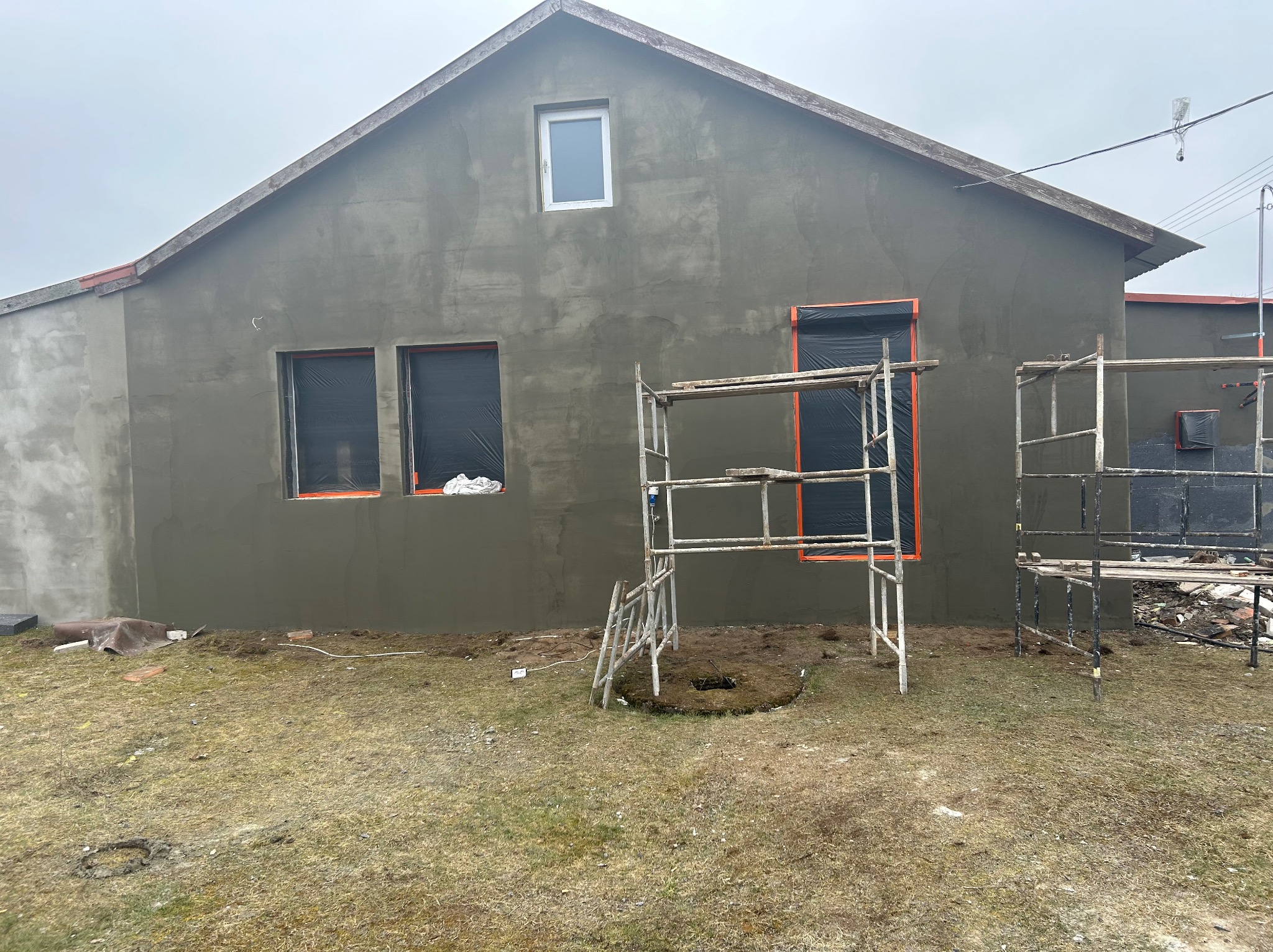 Dom w trakcie tynkowania elewacji, okna zabezpieczone folią, widoczne rusztowania na trawniku, surowy tynk w kolorze szarym.