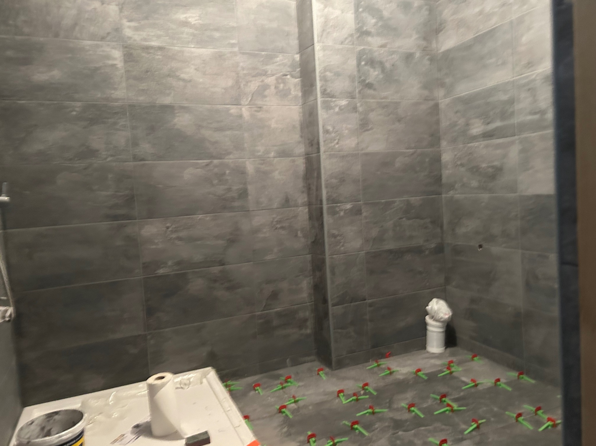 Świeżo ułożone, szare, prostokątne płytki w kabinie prysznicowej, widoczne krzyżyki dystansowe, wiadro z klejem i rolka papieru na wannie.