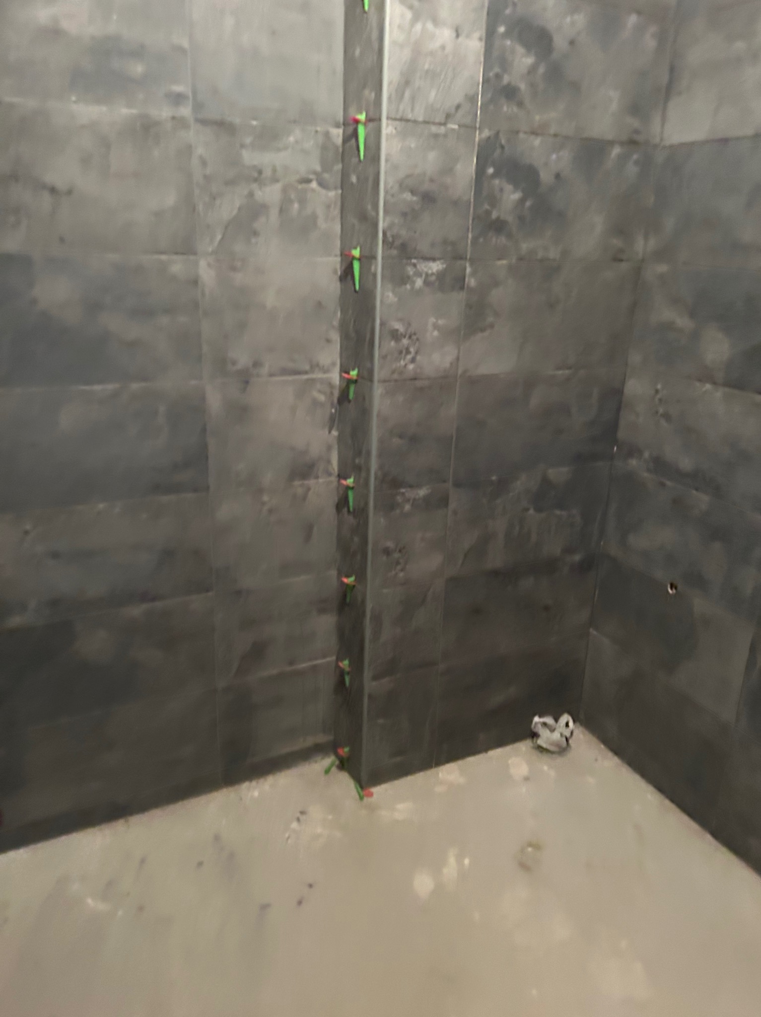 Układanie ciemnoszarych płytek na ścianie z użyciem klinów poziomujących, widoczny fragment podłogi z pozostałościami zaprawy.