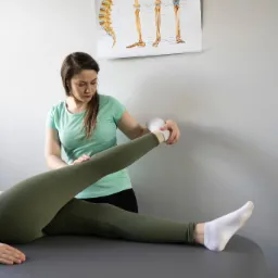 Kobieta w turkusowej koszulce wykonuje ćwiczenia rozciągające nogę pacjentki na szarym stole do rehabilitacji. W tle plakat z układem kostnym.