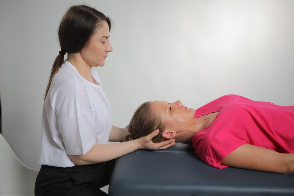 Kobieta w białej koszulce delikatnie masuje szyję pacjentki leżącej na niebieskim stole do masażu w jasnym gabinecie.