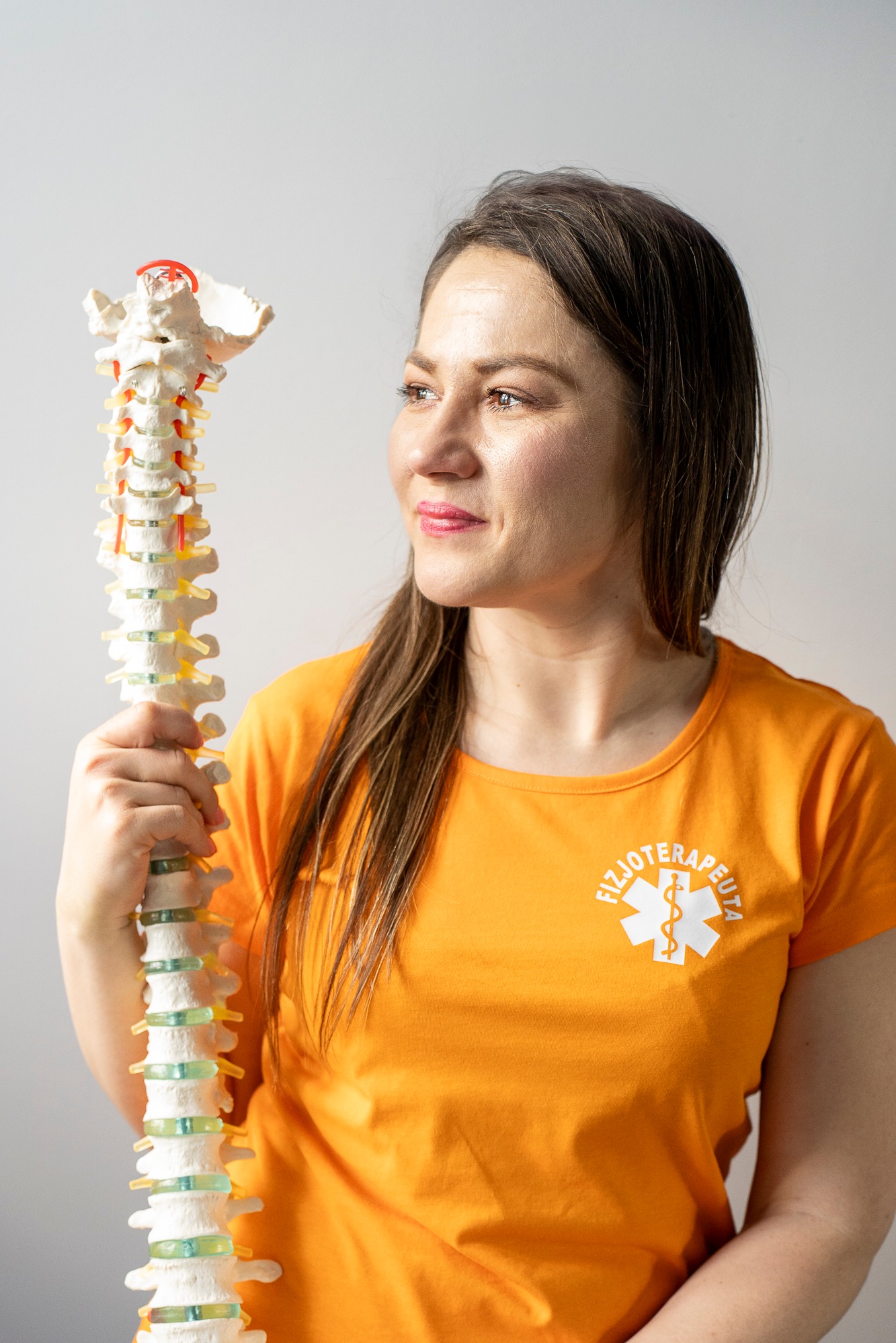 Kobieta w pomarańczowej koszulce z symbolem fizjoterapii trzyma model kręgosłupa, patrząc w prawo. Jasne tło, neutralny wyraz twarzy.