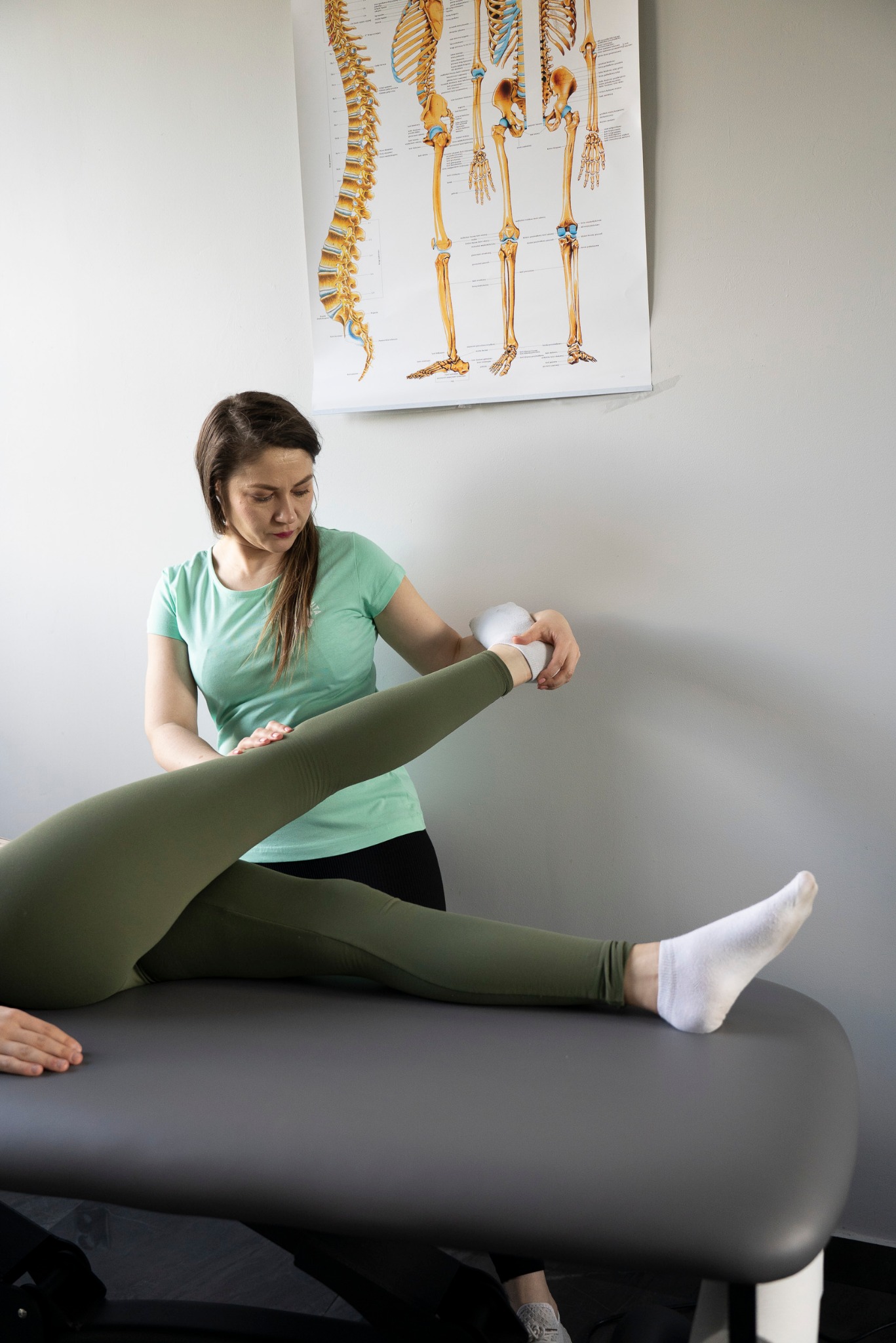 Kobieta w turkusowej koszulce wykonuje ćwiczenia rozciągające nogę pacjentki na szarym stole do rehabilitacji. W tle plakat z układem kostnym.
