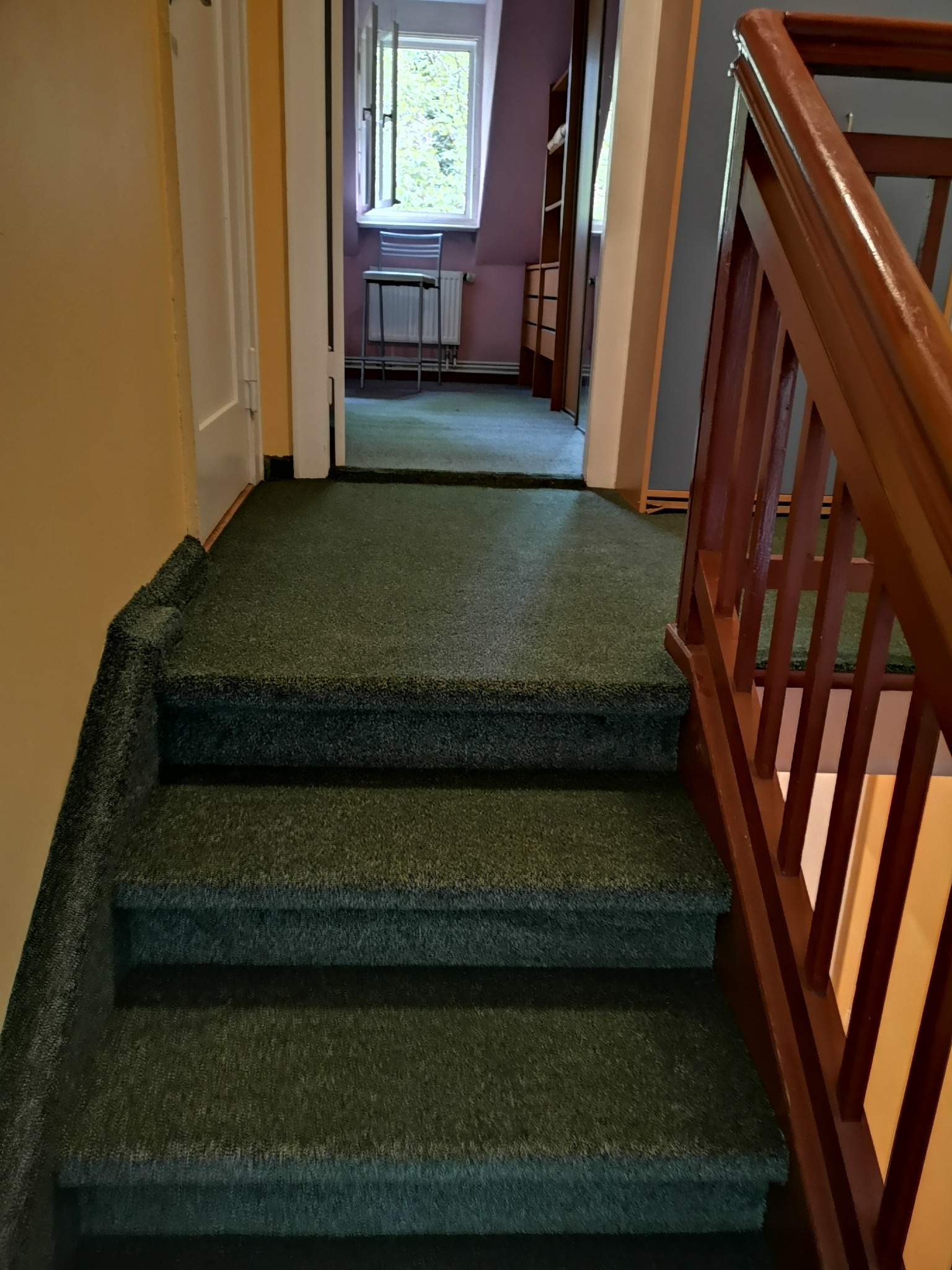 Schody pokryte zieloną wykładziną dywanową, widoczne drewniane poręcze i fragment korytarza z oknem w tle.