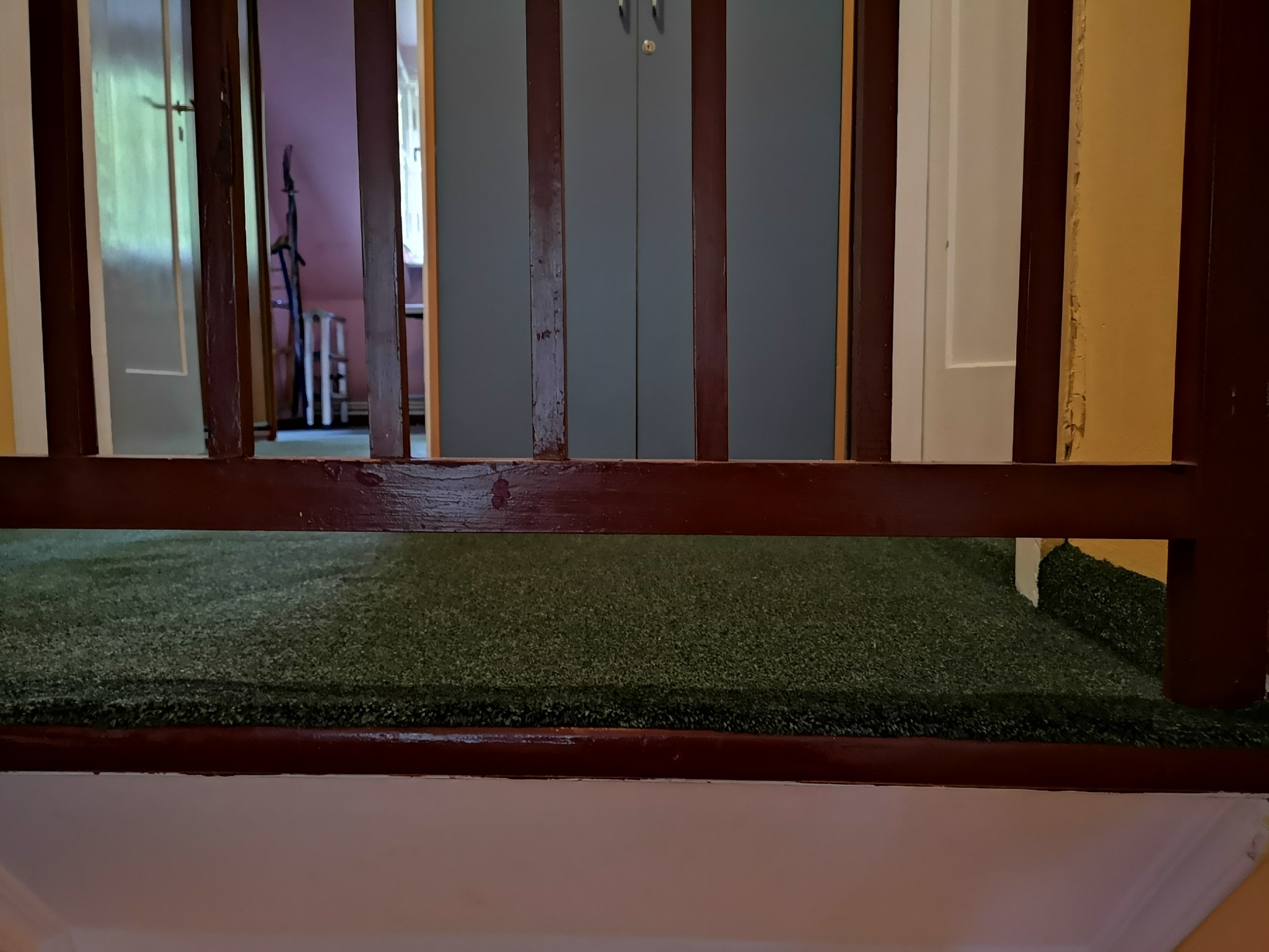 Fragment schodów z zieloną wykładziną dywanową imitującą trawę, widok przez drewnianą balustradę, w tle drzwi i szafa.