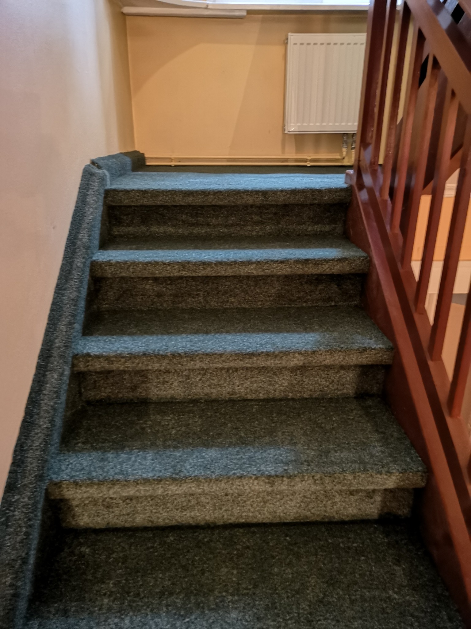 Schody pokryte szarozieloną wykładziną dywanową z drewnianą balustradą po prawej stronie, widoczny również grzejnik na ścianie.