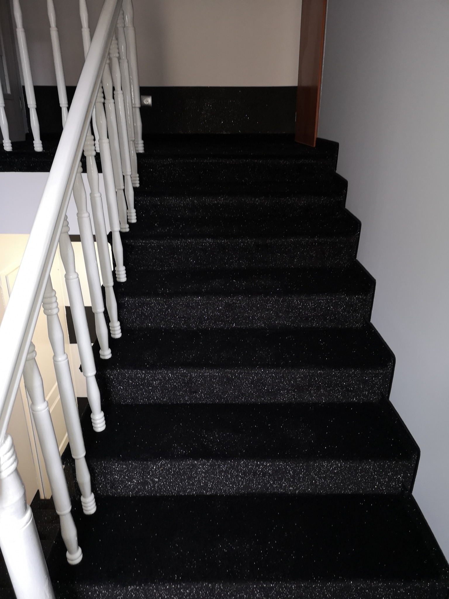 Czarne schody pokryte wykładziną dywanową z białymi balustradami i widocznym fragmentem drzwi w tle.