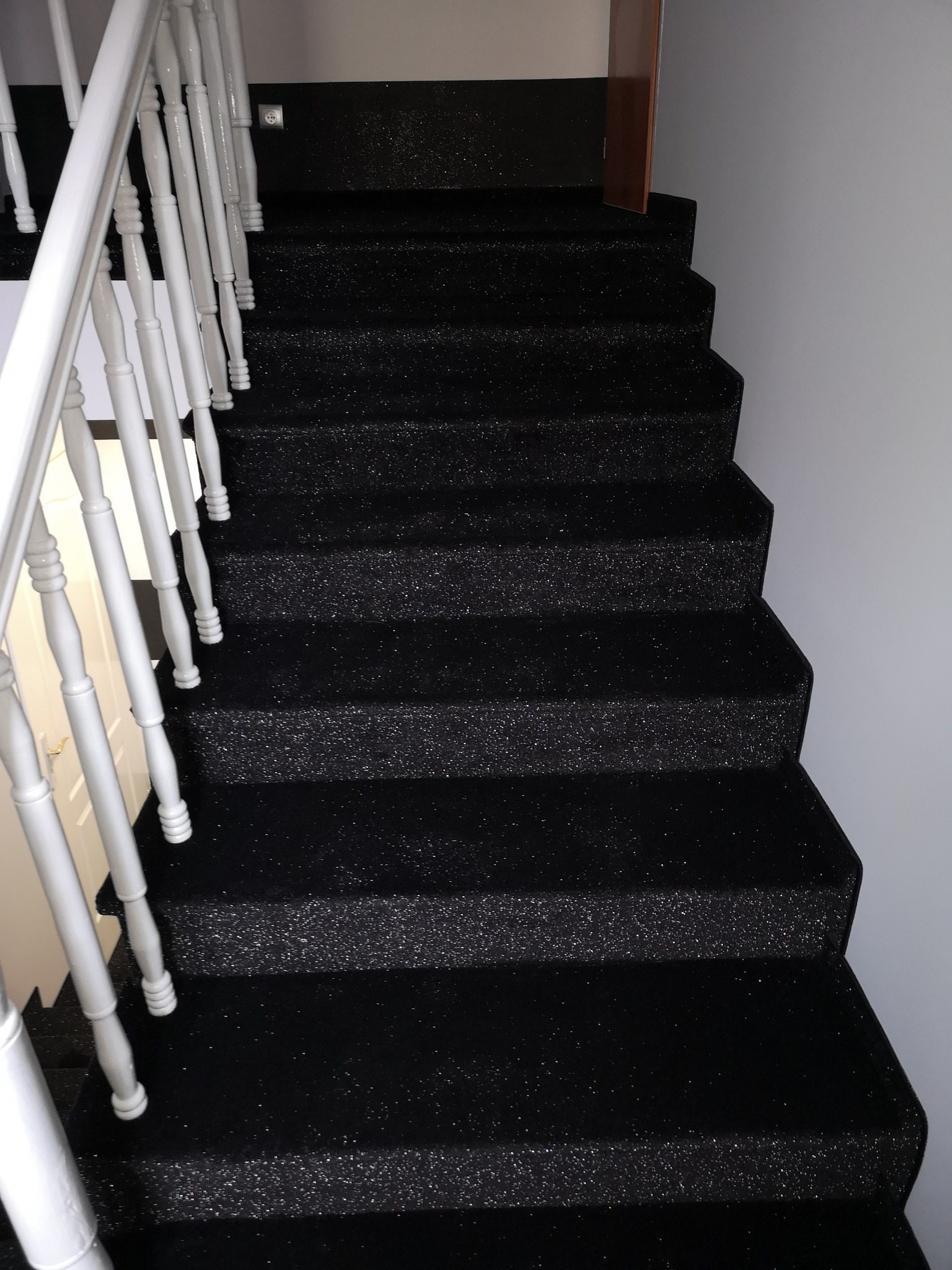Błyszcząca, czarna wykładzina dywanowa na schodach z białymi, drewnianymi poręczami i widocznym gniazdkiem elektrycznym na ścianie.