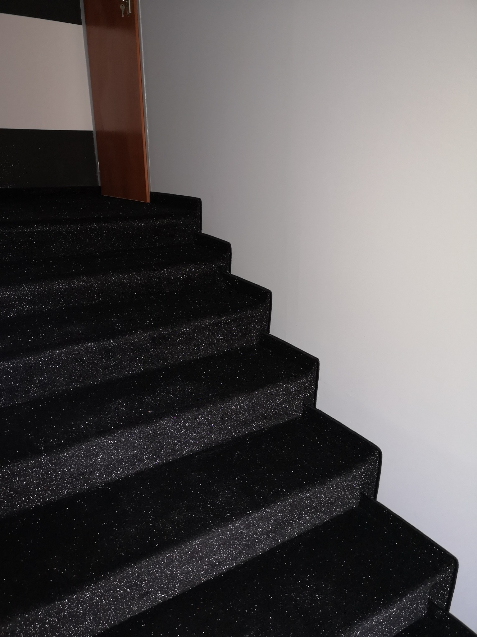 Schody pokryte czarną, błyszczącą wykładziną dywanową, widoczne fragmenty białej ściany i brązowych drzwi.
