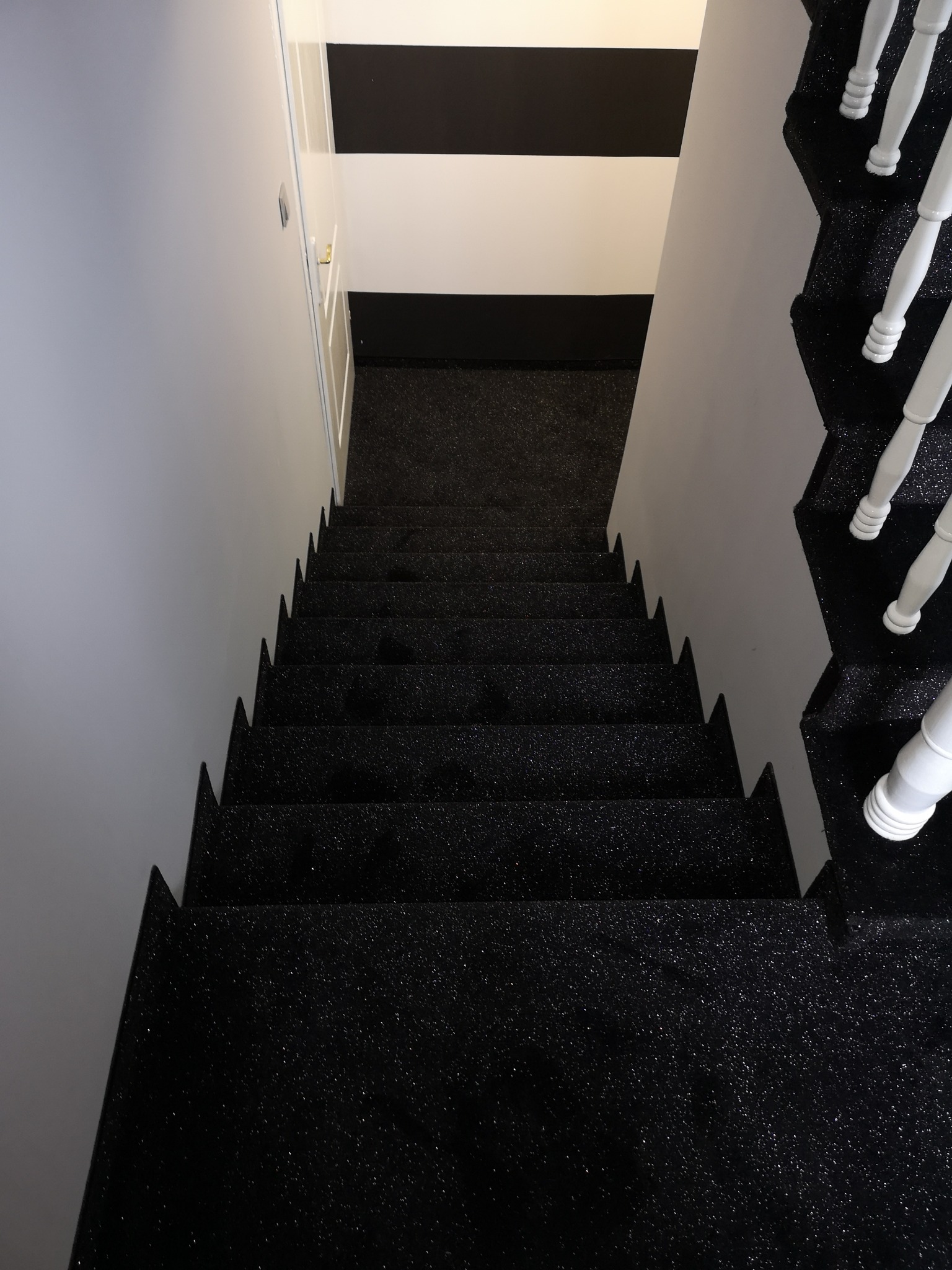 Schody pokryte czarną, błyszczącą wykładziną, widok z góry, białe poręcze, ściana z czarno-białymi pasami w tle.
