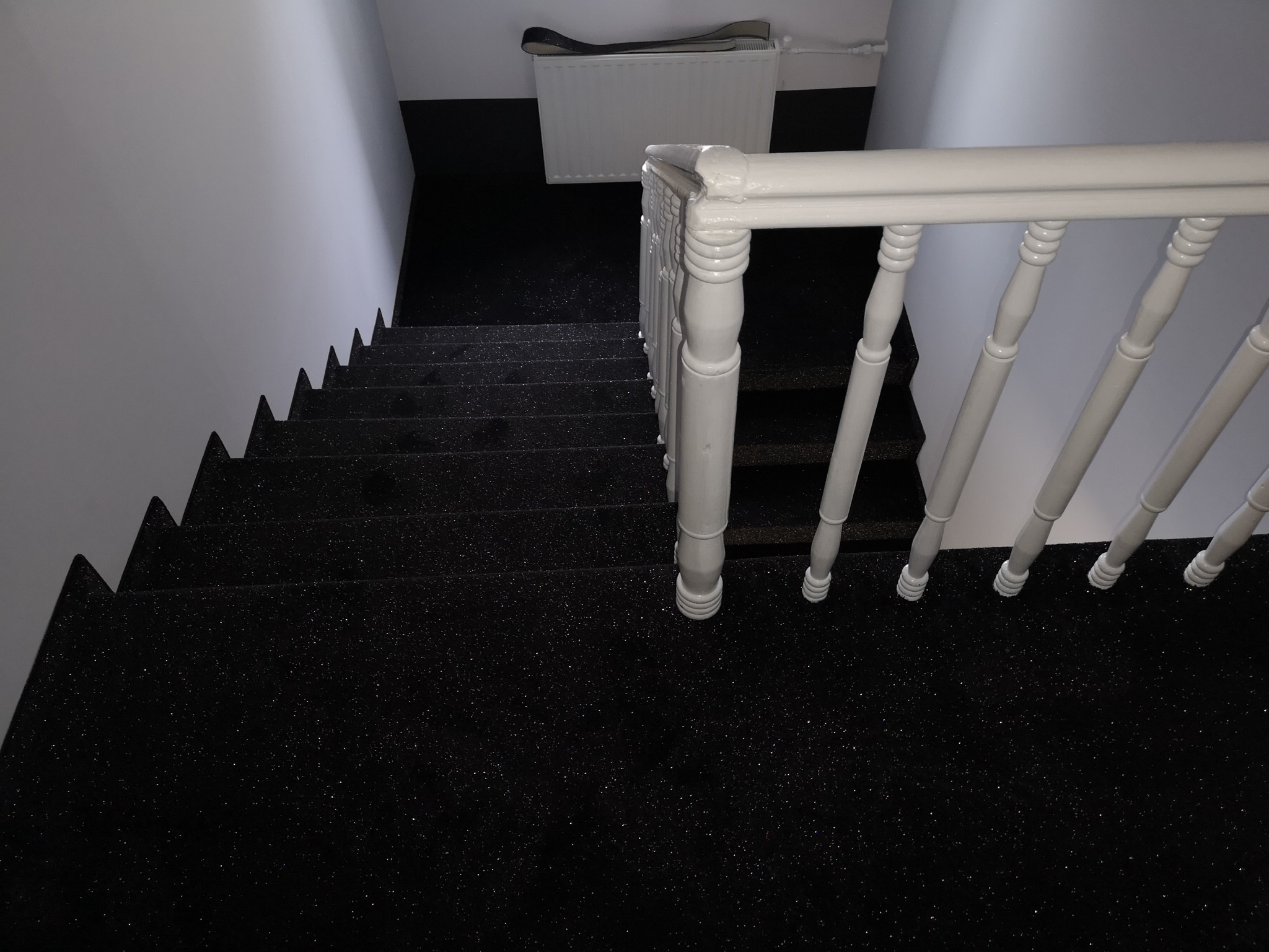 Widok z góry na schody pokryte czarną, błyszczącą wykładziną dywanową, z białymi balustradami i grzejnikiem w tle.