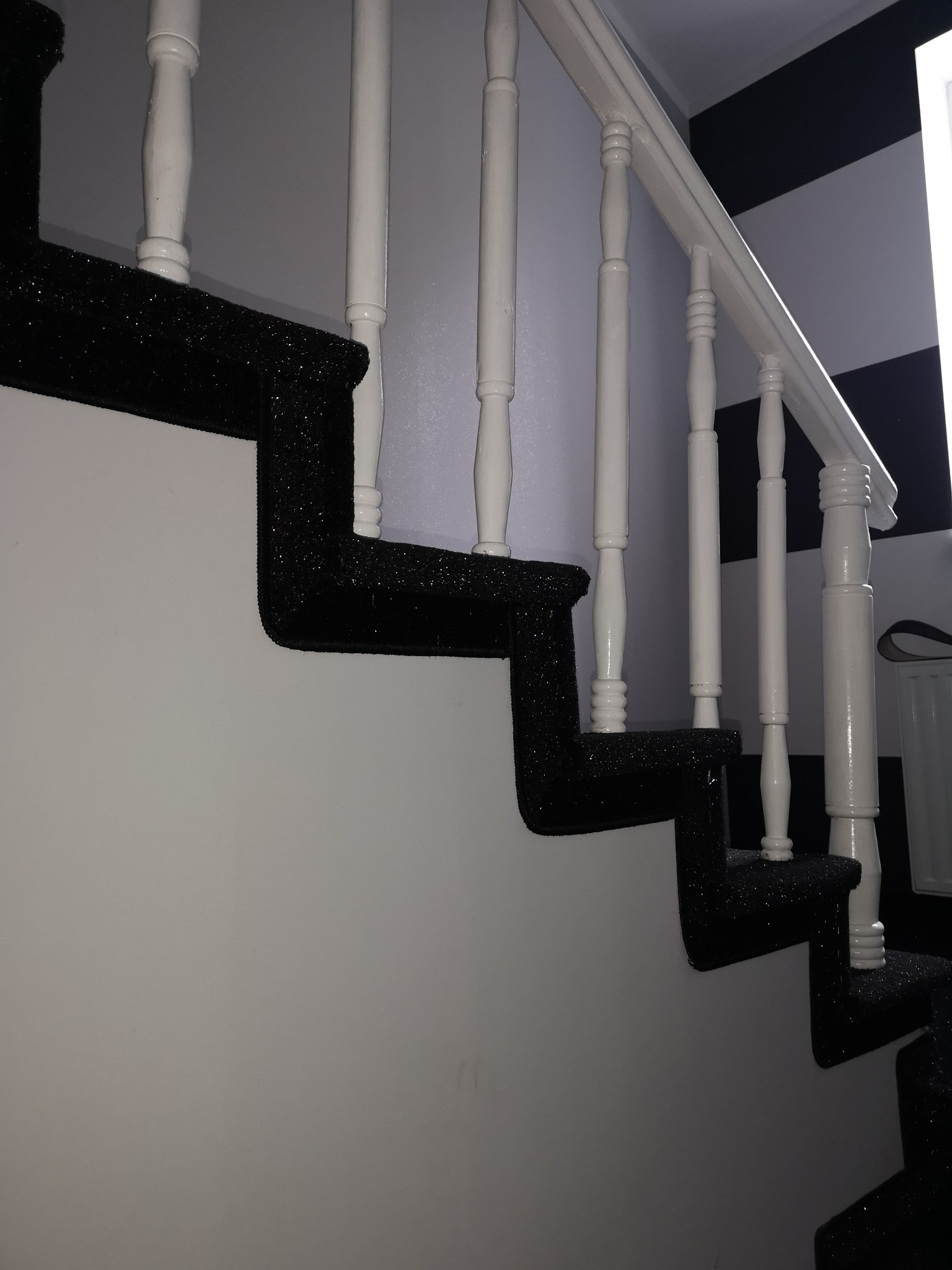 Schody obite czarną, brokatową wykładziną dywanową, z białymi, drewnianymi balustradami na tle ściany w czarno-białe pasy.