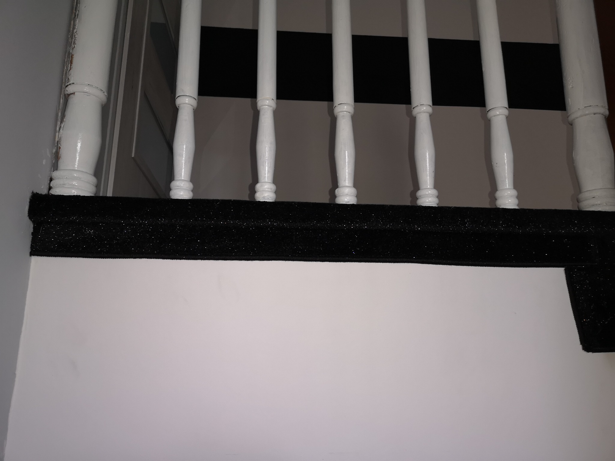 Fragment schodów z białymi balustradami i czarną wykładziną na stopniu, widoczne ślady zużycia na farbie balustrady.