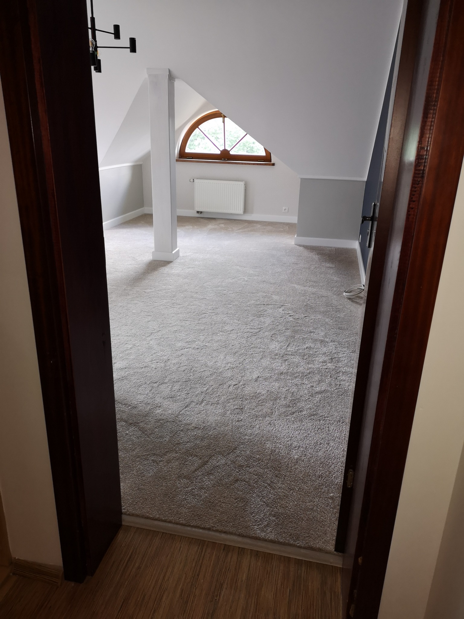 Jasnoszara wykładzina dywanowa w pokoju na poddaszu z oknem w kształcie półkola, widok zza drewnianych drzwi.