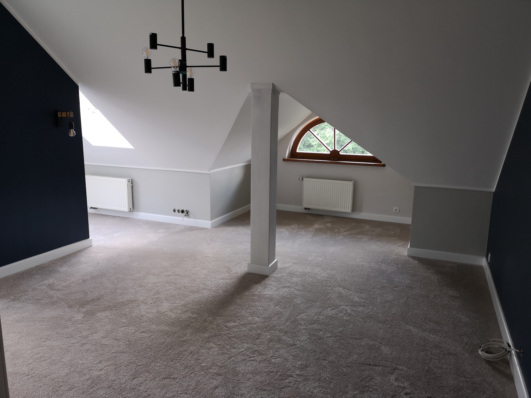 Przestronne poddasze po remoncie z beżową wykładziną dywanową, kolumną wspierającą i nowoczesnym żyrandolem.