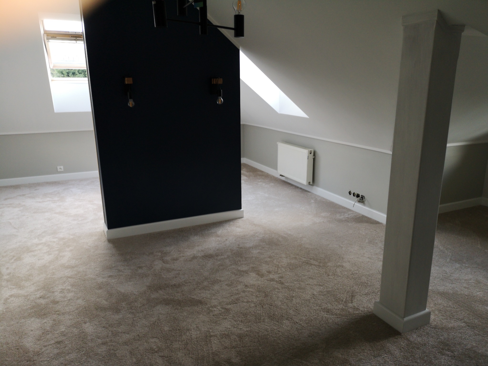 Przestronne poddasze z beżową wykładziną dywanową, widok na czarną ścianę z oświetleniem i okno dachowe.
