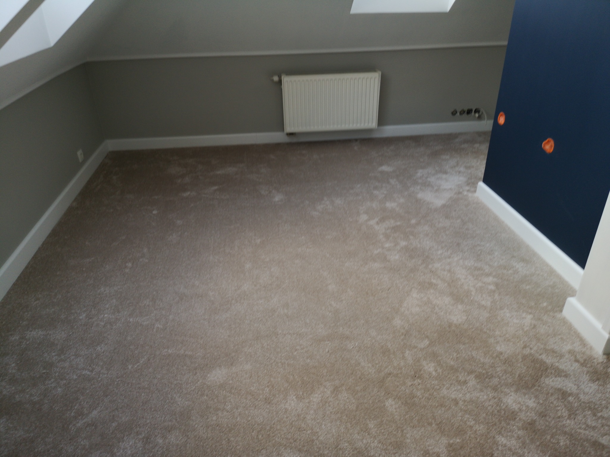 Beżowa wykładzina dywanowa w pokoju na poddaszu, białe listwy przypodłogowe, szare ściany z grzejnikiem i fragment granatowej ściany.