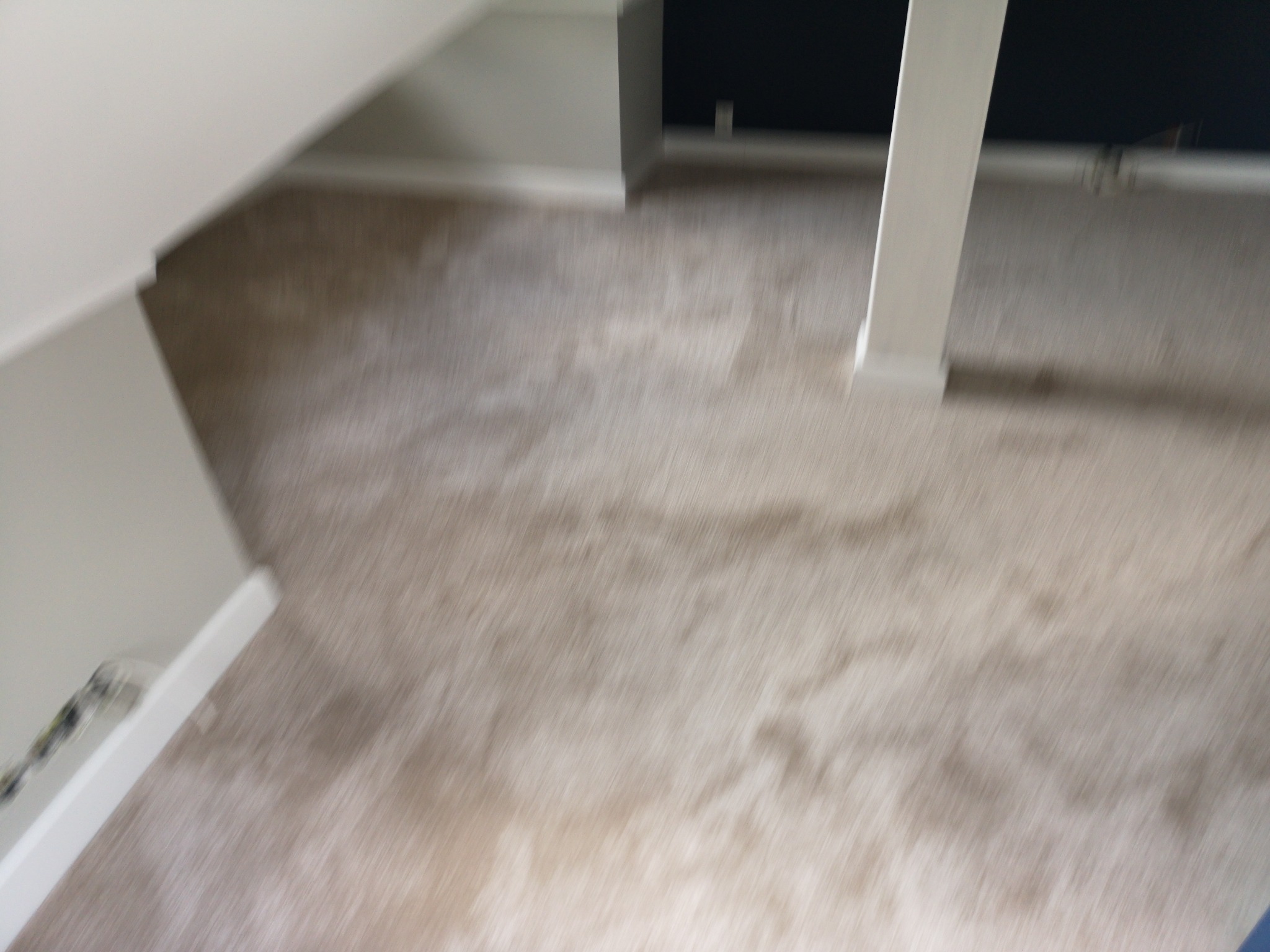 Beżowa wykładzina dywanowa w pokoju, widoczny fragment ściany i biała kolumna, zdjęcie z góry z lekkim rozmyciem.