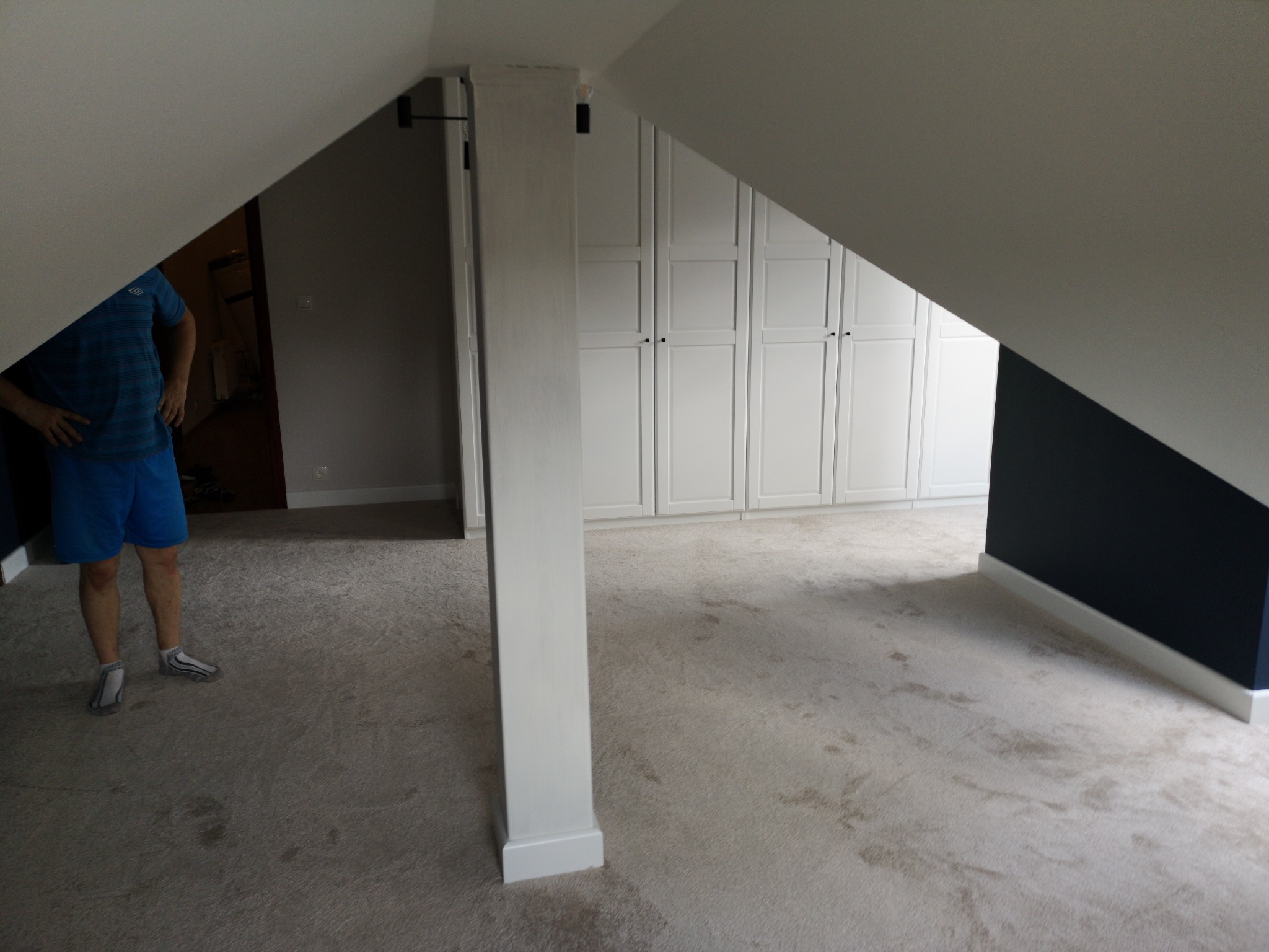 Pokój na poddaszu z beżową wykładziną dywanową, białą zabudową meblową pod skosem i fragmentem osoby w niebieskim stroju sportowym.