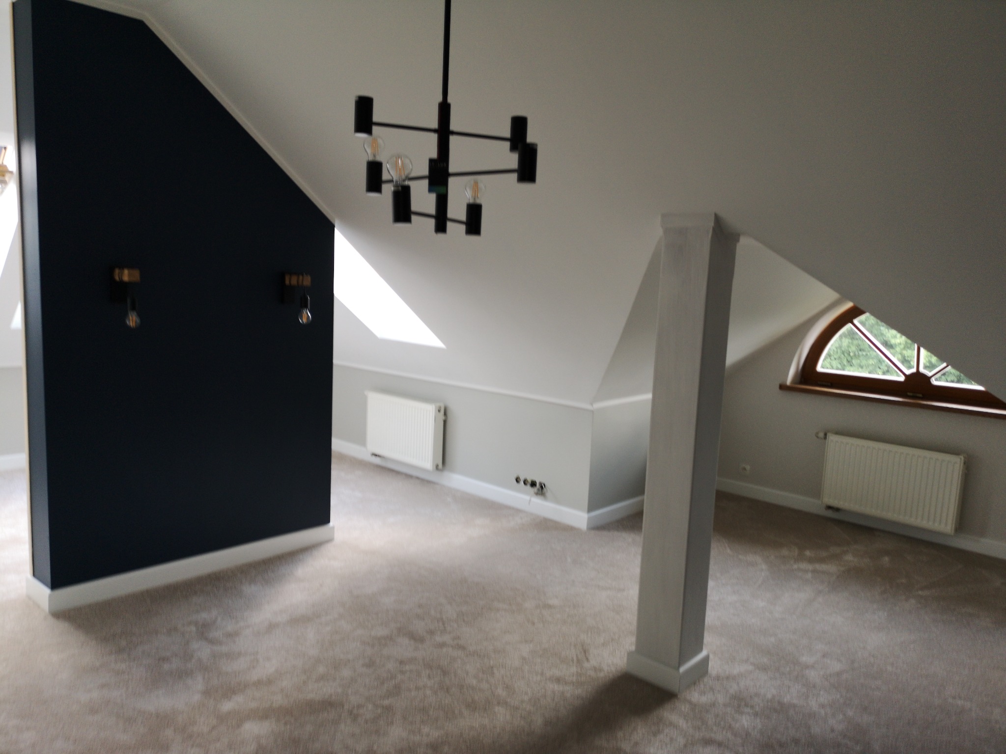 Nowoczesne poddasze z beżową wykładziną dywanową, czarną ścianą akcentującą i designerską lampą sufitową.