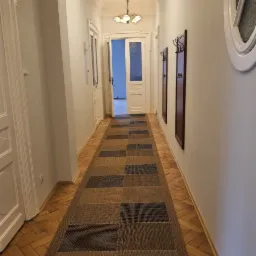 Długi korytarz w kamienicy z parkietem w jodełkę i dywanem w brązowo-czarne kwadraty. Na ścianie wieszaki z ciemnego drewna, okrągłe okno i drzwi wejściowe.